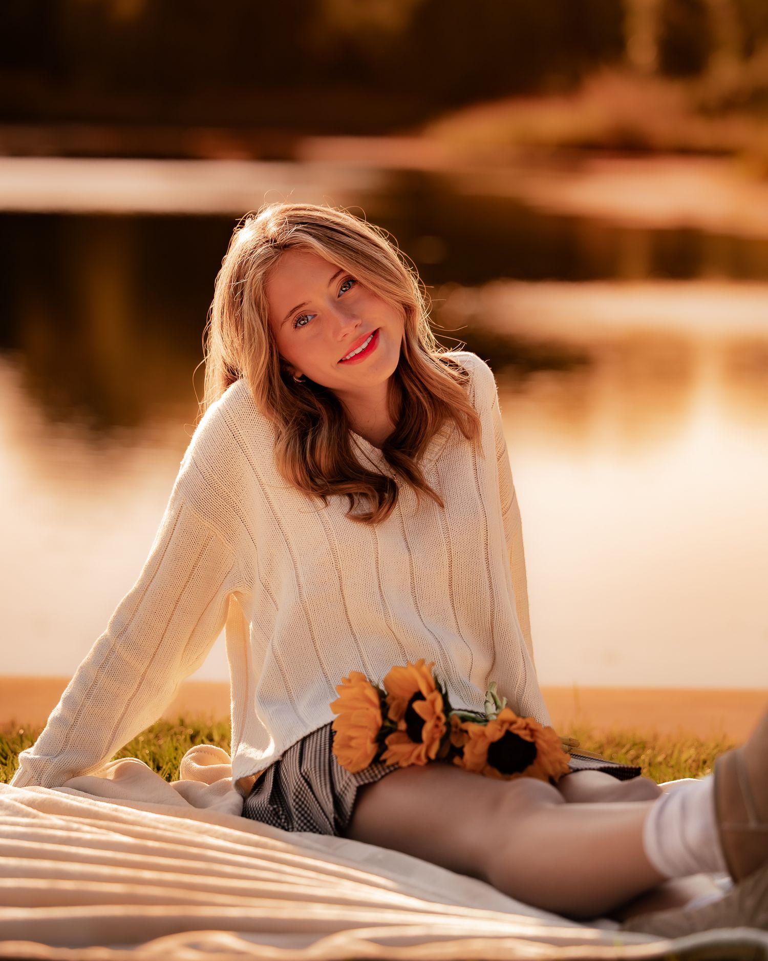 Young woman in cream sweater holding sunflowers, sitting on blanket by golden lake at sunset.