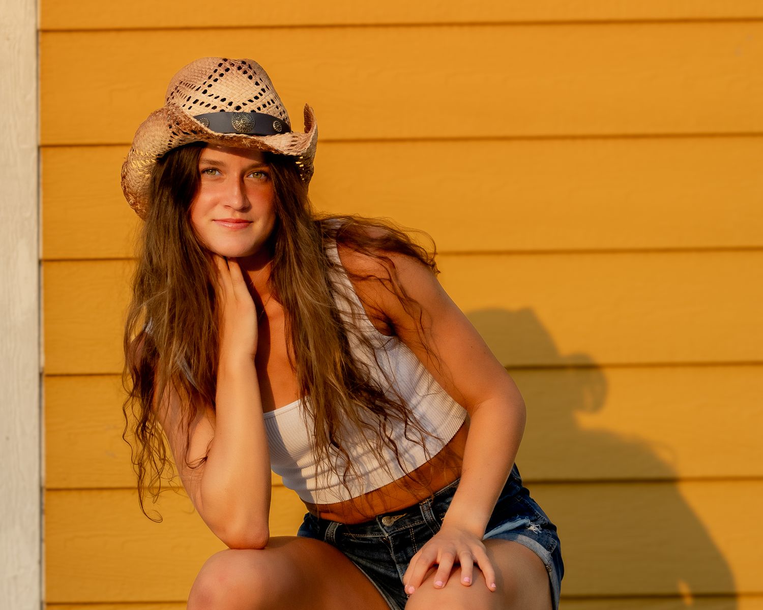 A person in a cowboy hat and denim shorts poses against a yellow wall in warm sunlight.