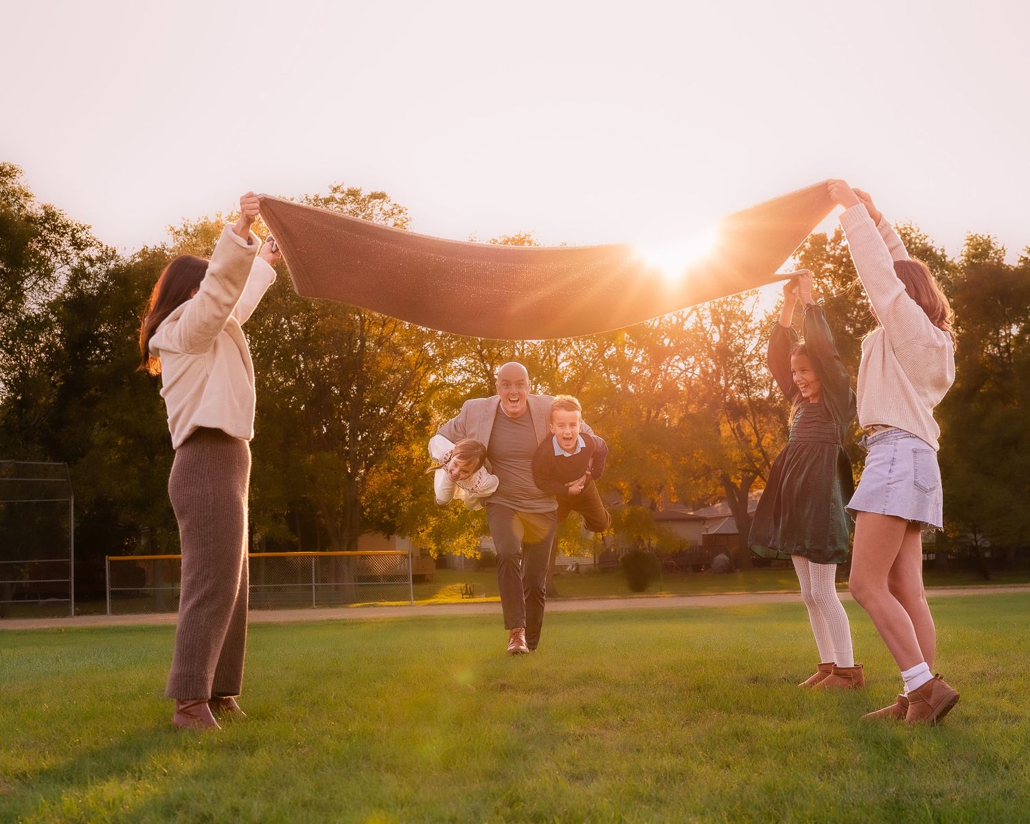 Family plays under blanket at sunset in park setting.