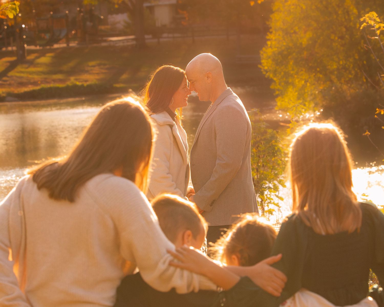 Couple sharing romantic moment by sunlit lake surrounded by family at golden hour.