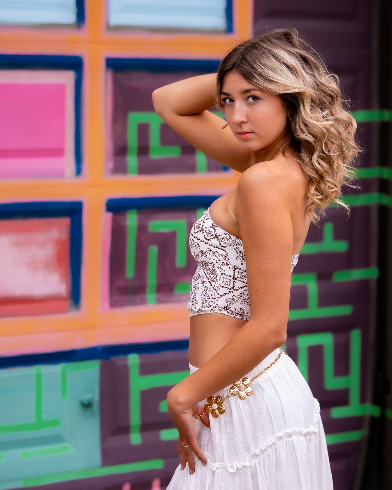 Young woman in white crop top and skirt poses confidently against colorful graffiti wall.