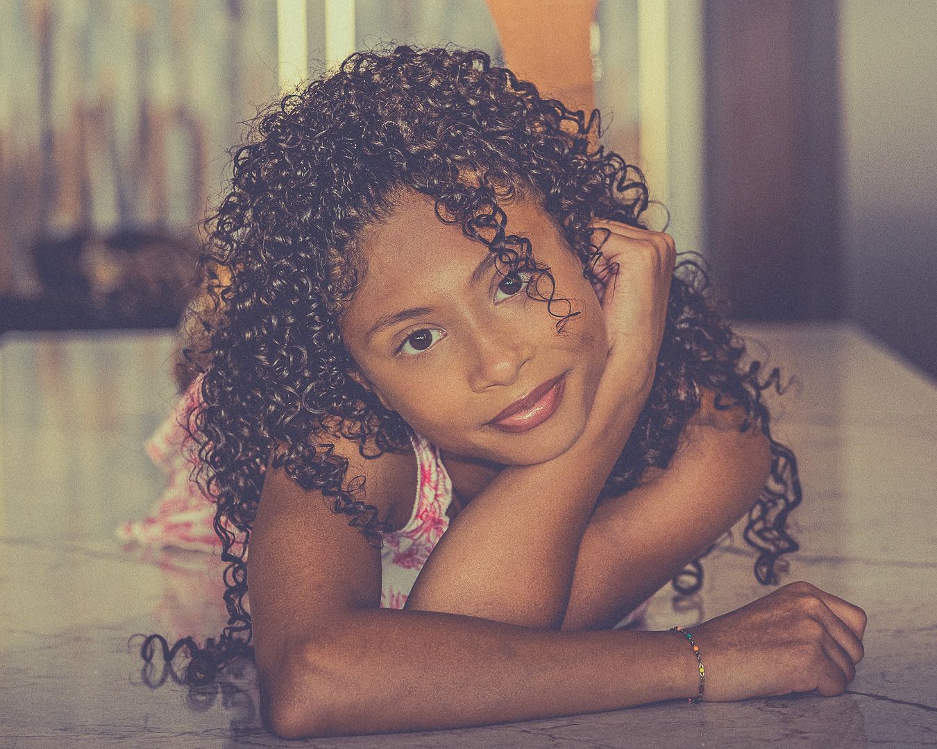 Young girl with curly hair lying on a surface, resting her chin on her hands, smiling softly at the camera.