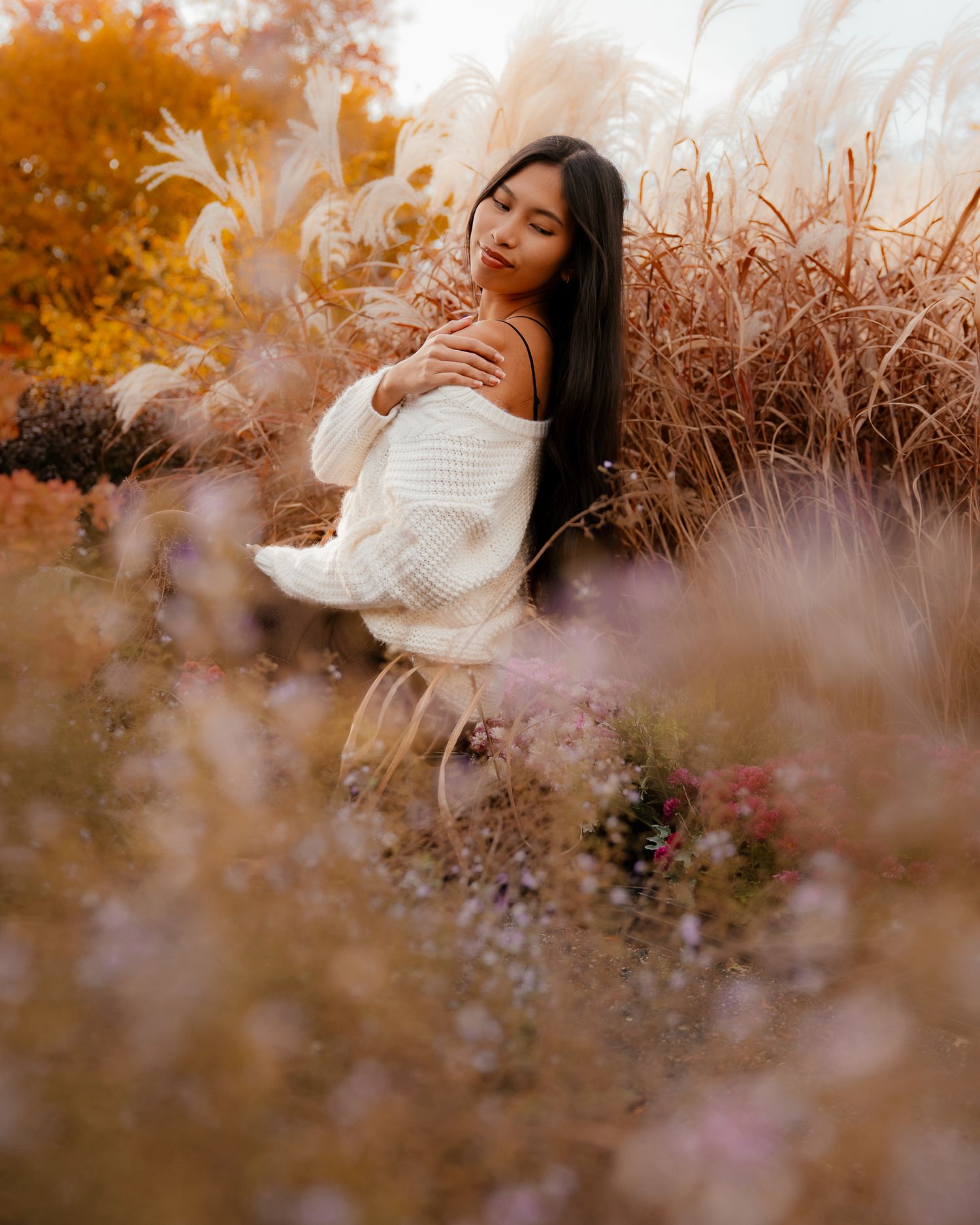 A person in a white sweater poses among autumn foliage and soft hazy lighting creating a dreamy outdoor portrait.