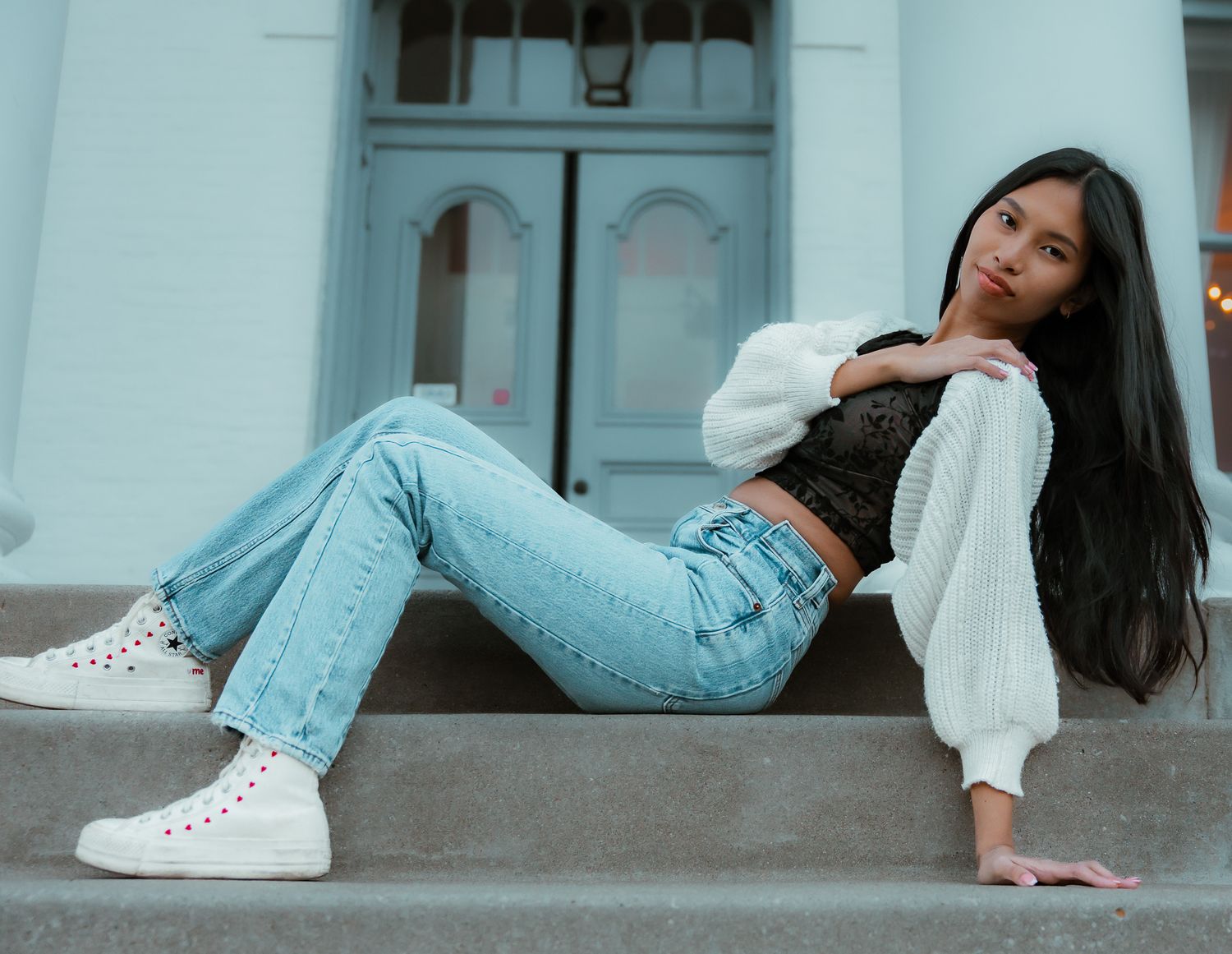 Casual street style look with light wash jeans, white cardigan, crop top and high-top sneakers on concrete steps.