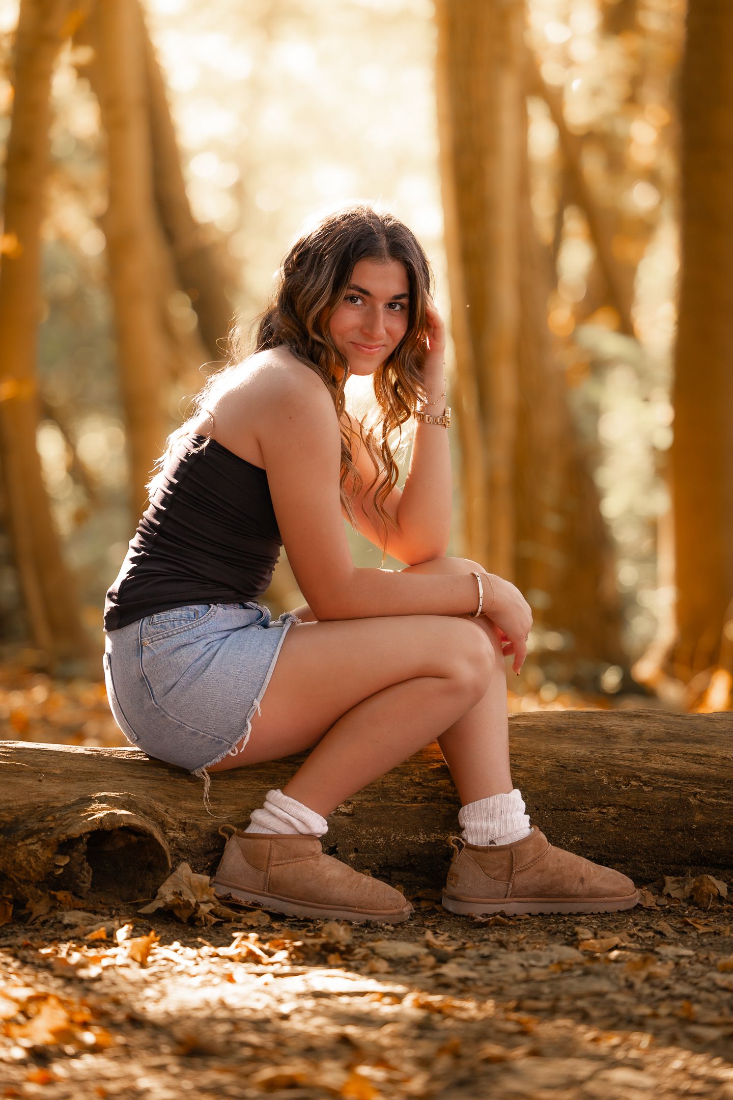 Young woman in denim shorts and UGG boots sits on a log in a sunlit autumn forest, smiling at the camera.