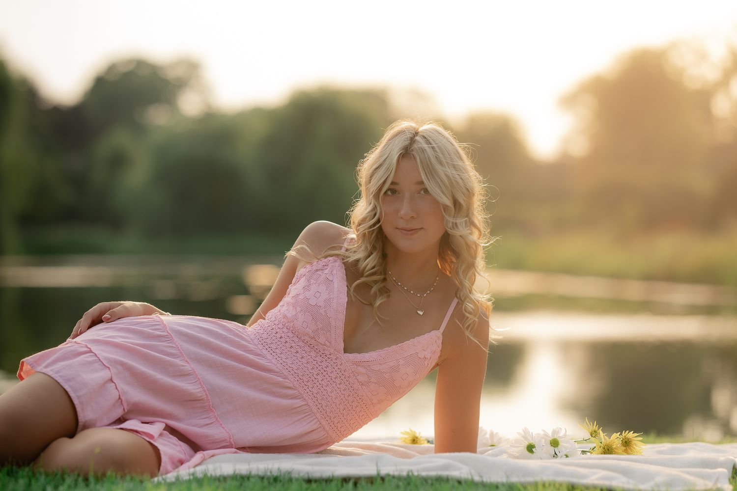 Woman in pink dress poses outdoors during golden hour sunset by a lake with dreamy lighting.