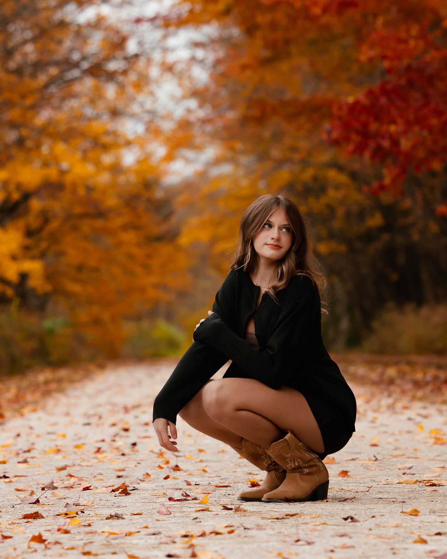 A person in a black sweater poses gracefully among fallen autumn leaves in a park with vibrant orange foliage.