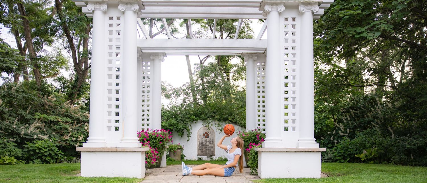 A white decorative garden archway with floral arrangements stands in a lush green outdoor setting.