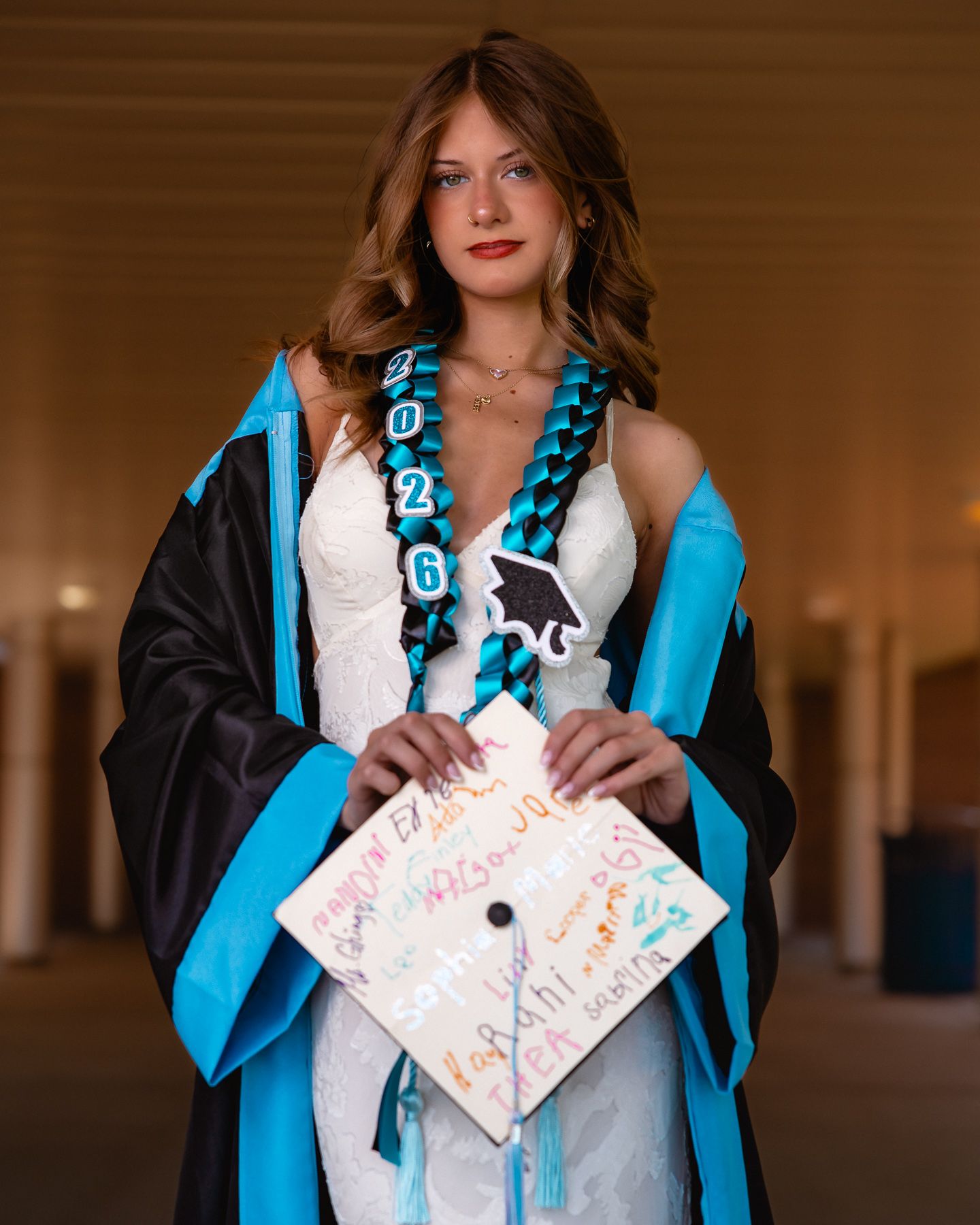 Young woman in teal and black graduation gown holding decorated cap with class of 2026 sash.