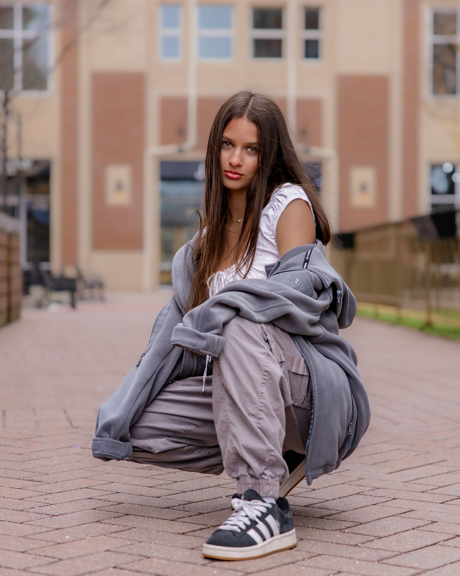 Young woman in gray cargo pants, white top, and Adidas sneakers crouching on brick pavement outdoors.