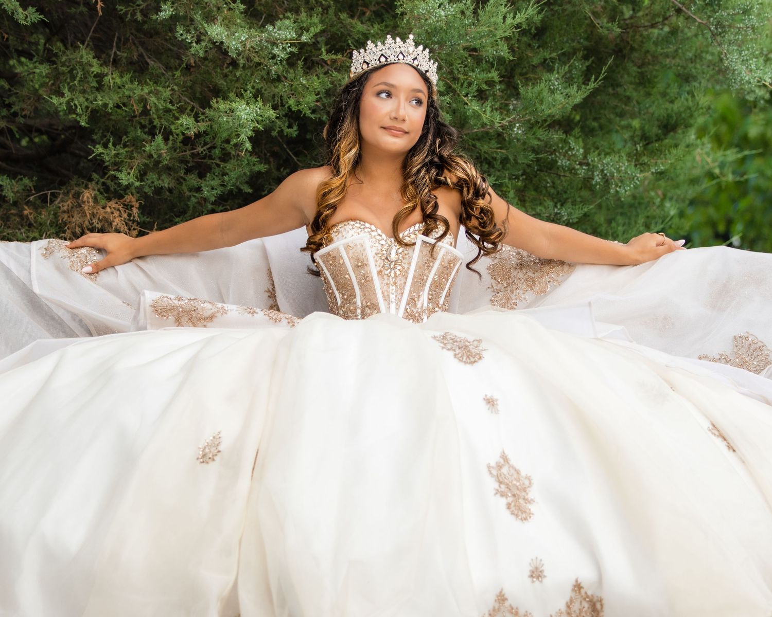 A stunning white quinceanera ball gown with delicate floral appliques and a beaded corset shown amidst green foliage.