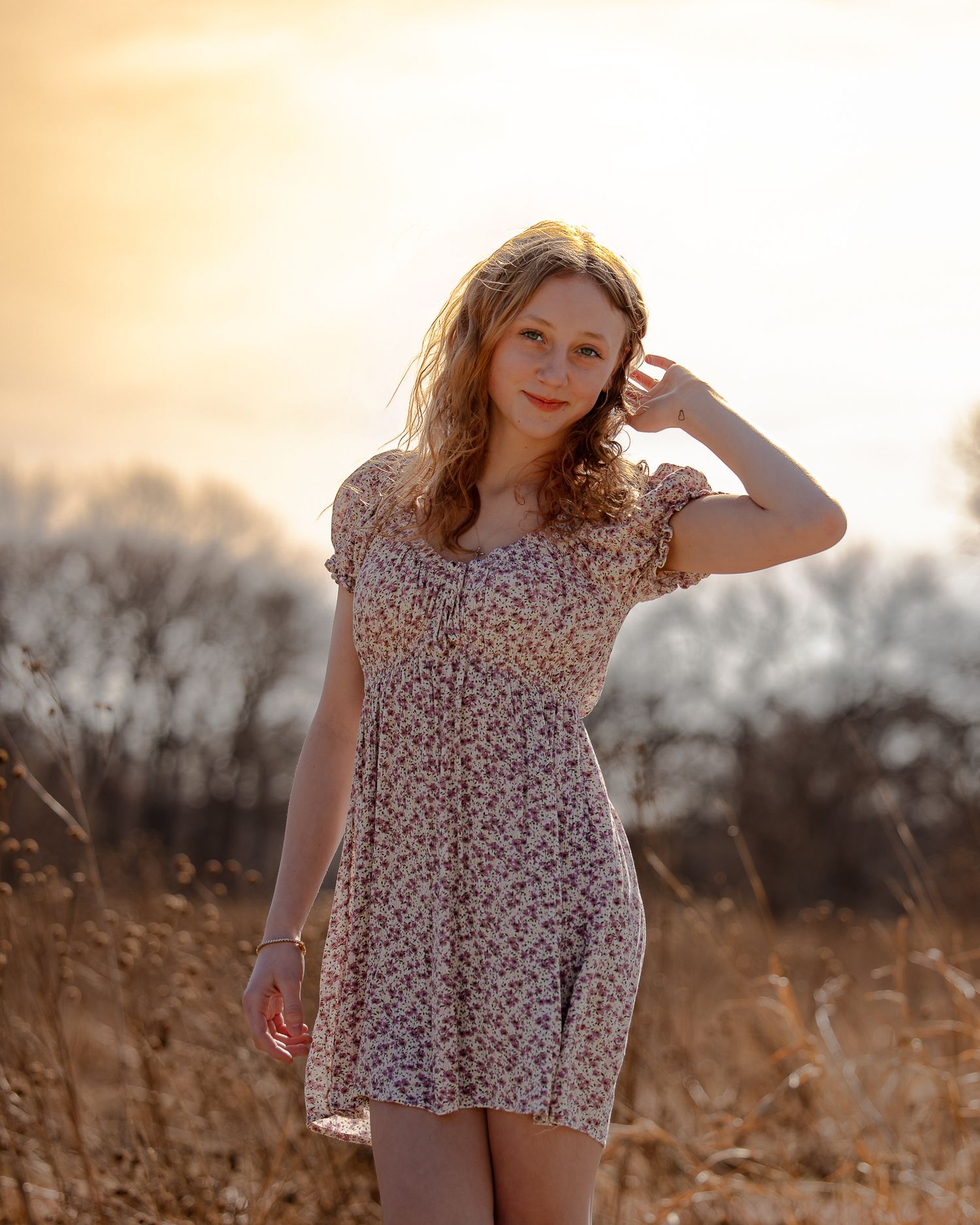 Young woman in floral sundress poses in golden field at sunset, hand in wavy hair, warm light glowing behind her.