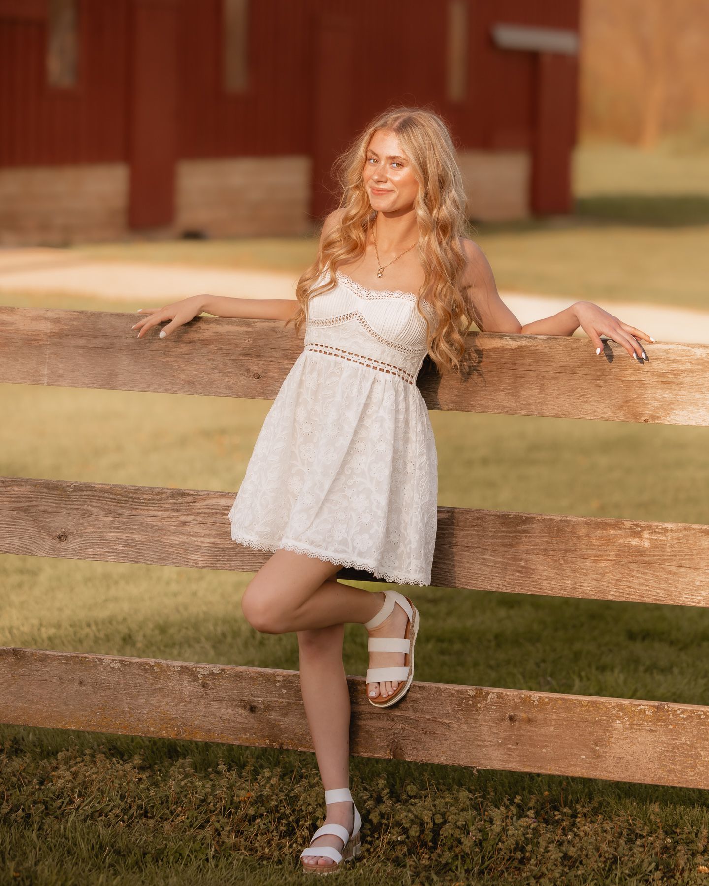 Young woman in white lace dress posing by a wooden fence at golden hour near a red barn.
