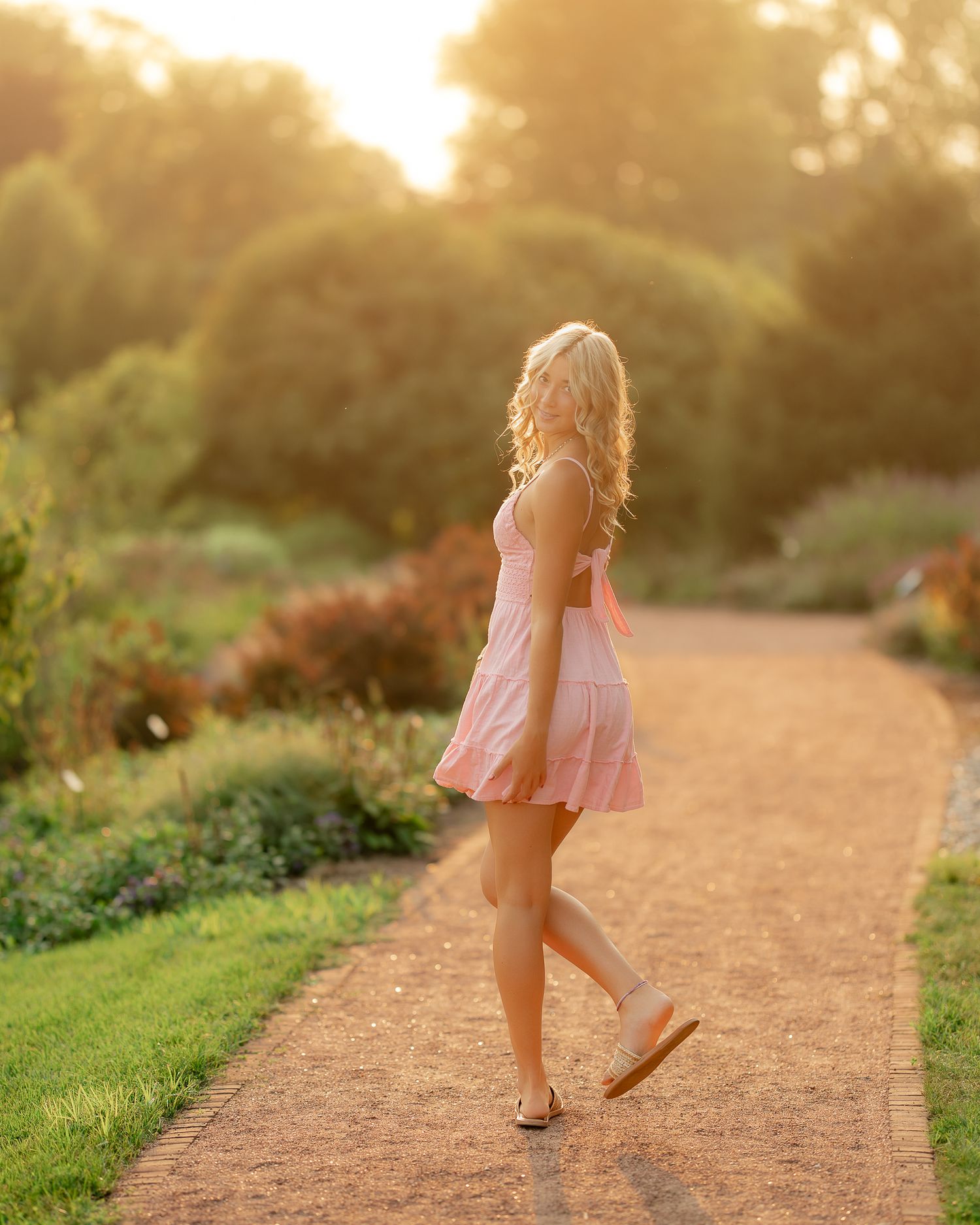 Blonde woman in pink sundress walking on garden path at golden hour sunset.