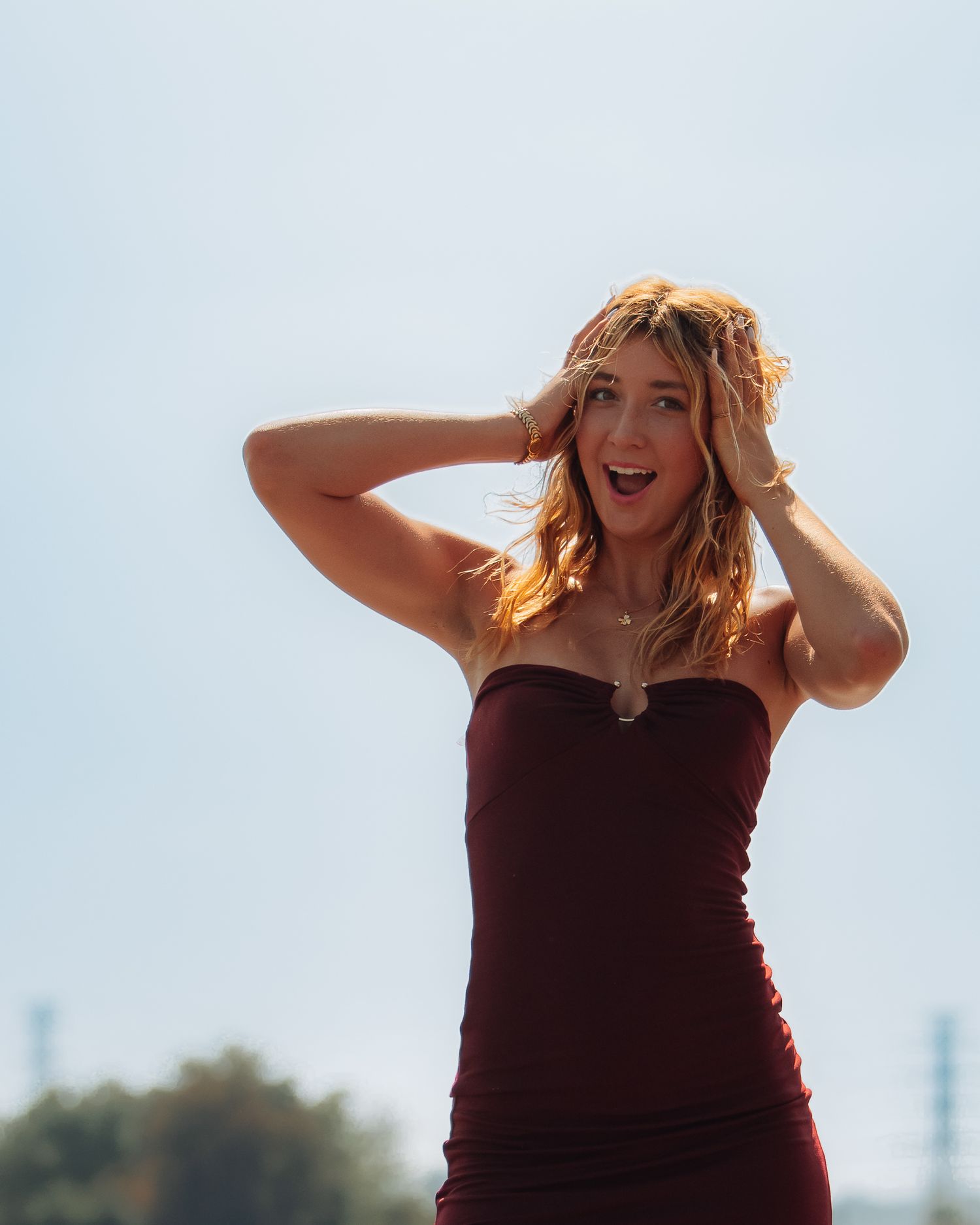 Person in burgundy dress poses expressively against bright sky background.