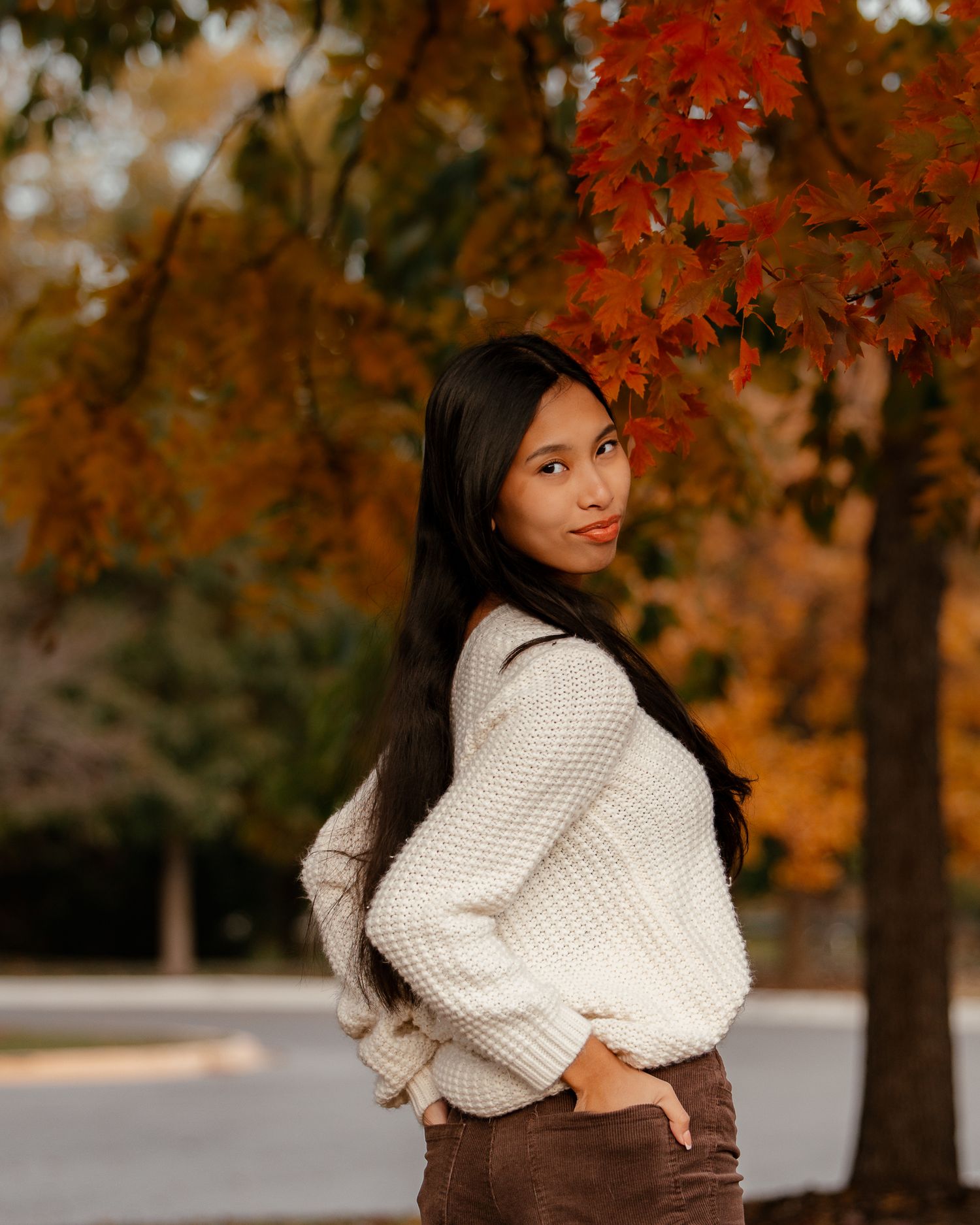 Person in cream sweater posing under autumn trees with orange leaves.