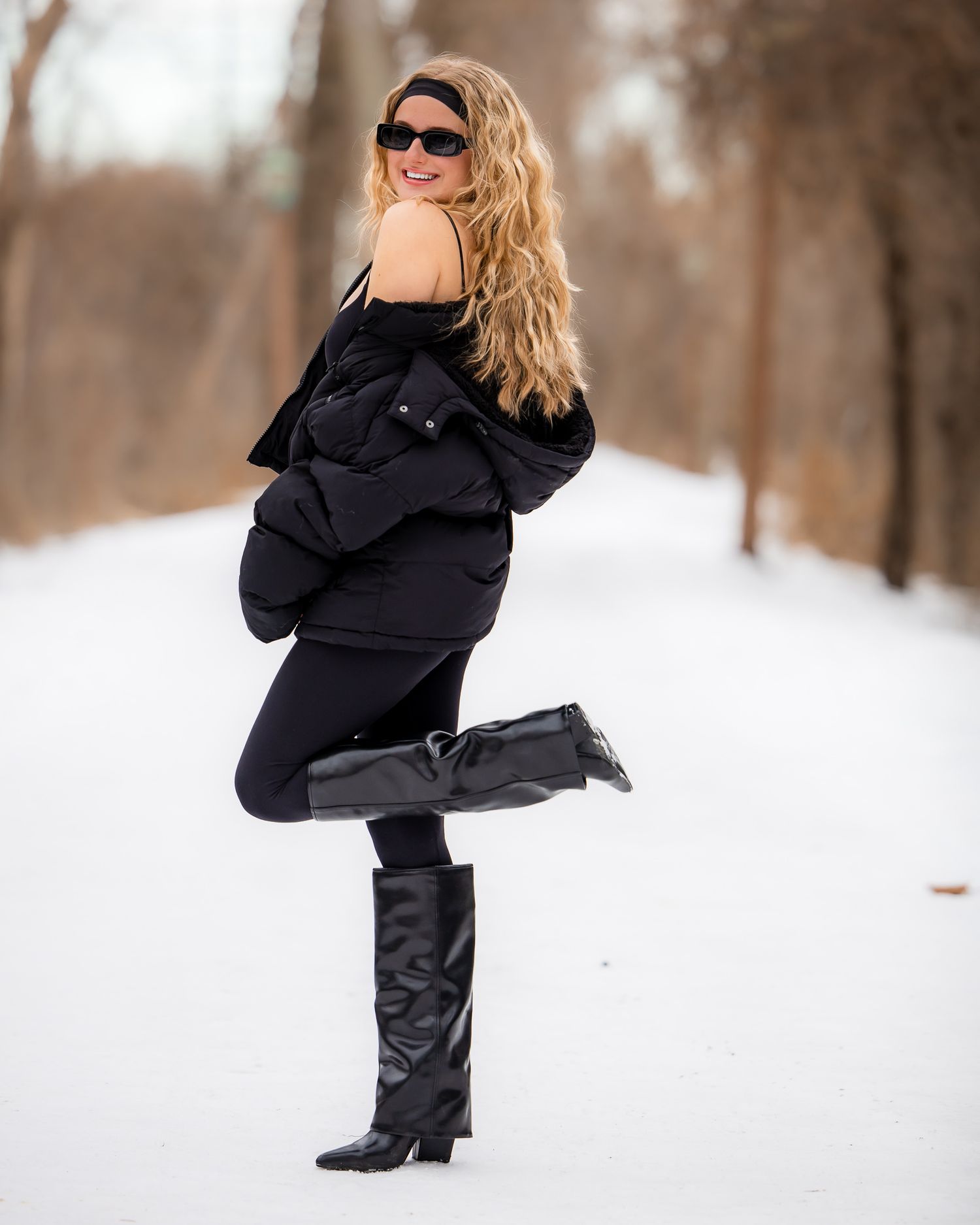 Stylish woman in black winter outfit with sunglasses poses playfully in snowy outdoor setting.