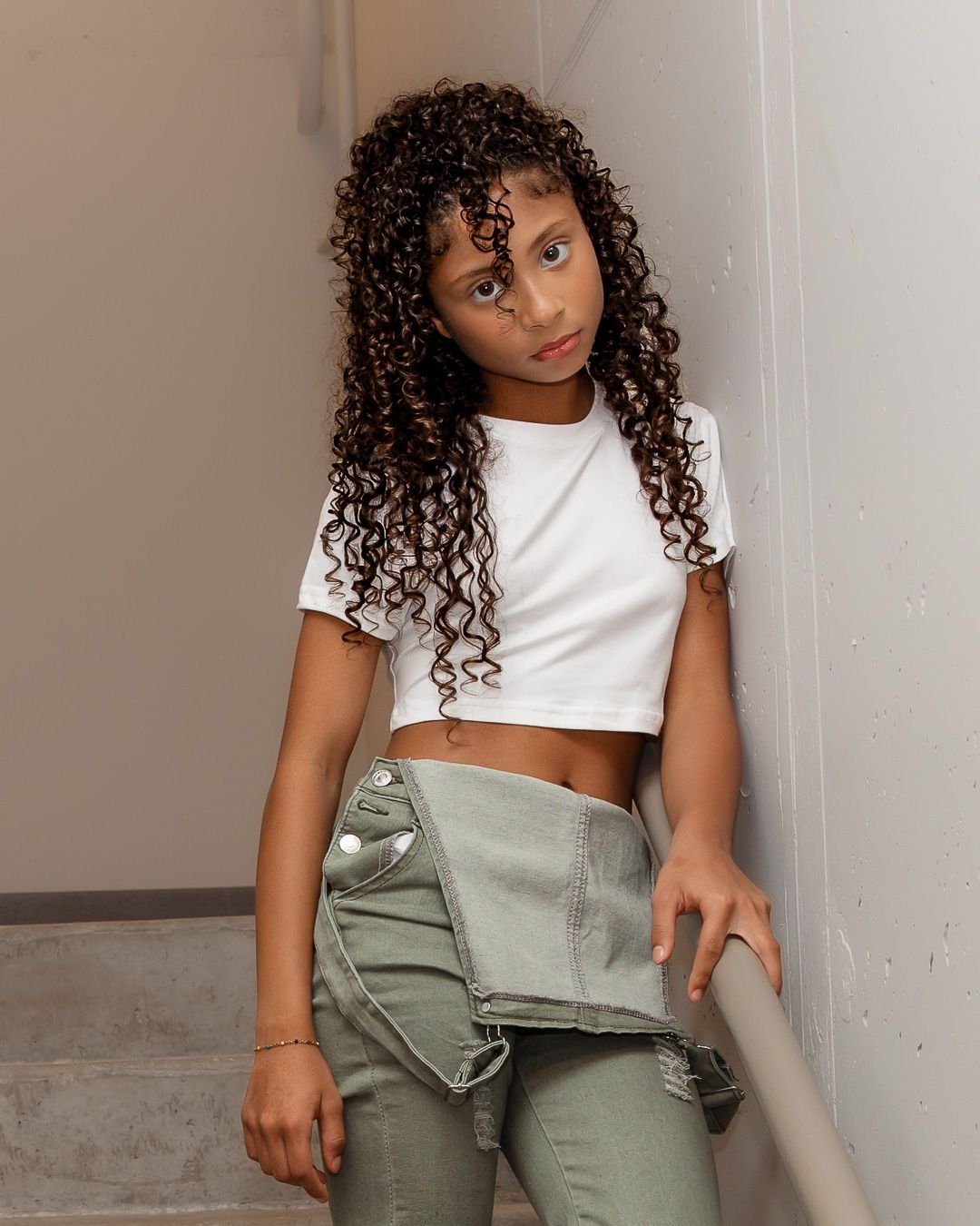 Young woman in white crop top and sage green jeans leans against wall on staircase.