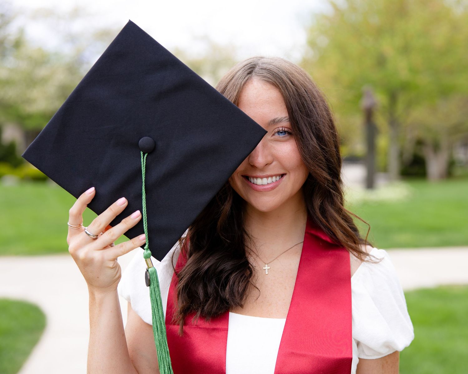 Graduate holds graduation cap while wearing red graduation stole outdoors.
