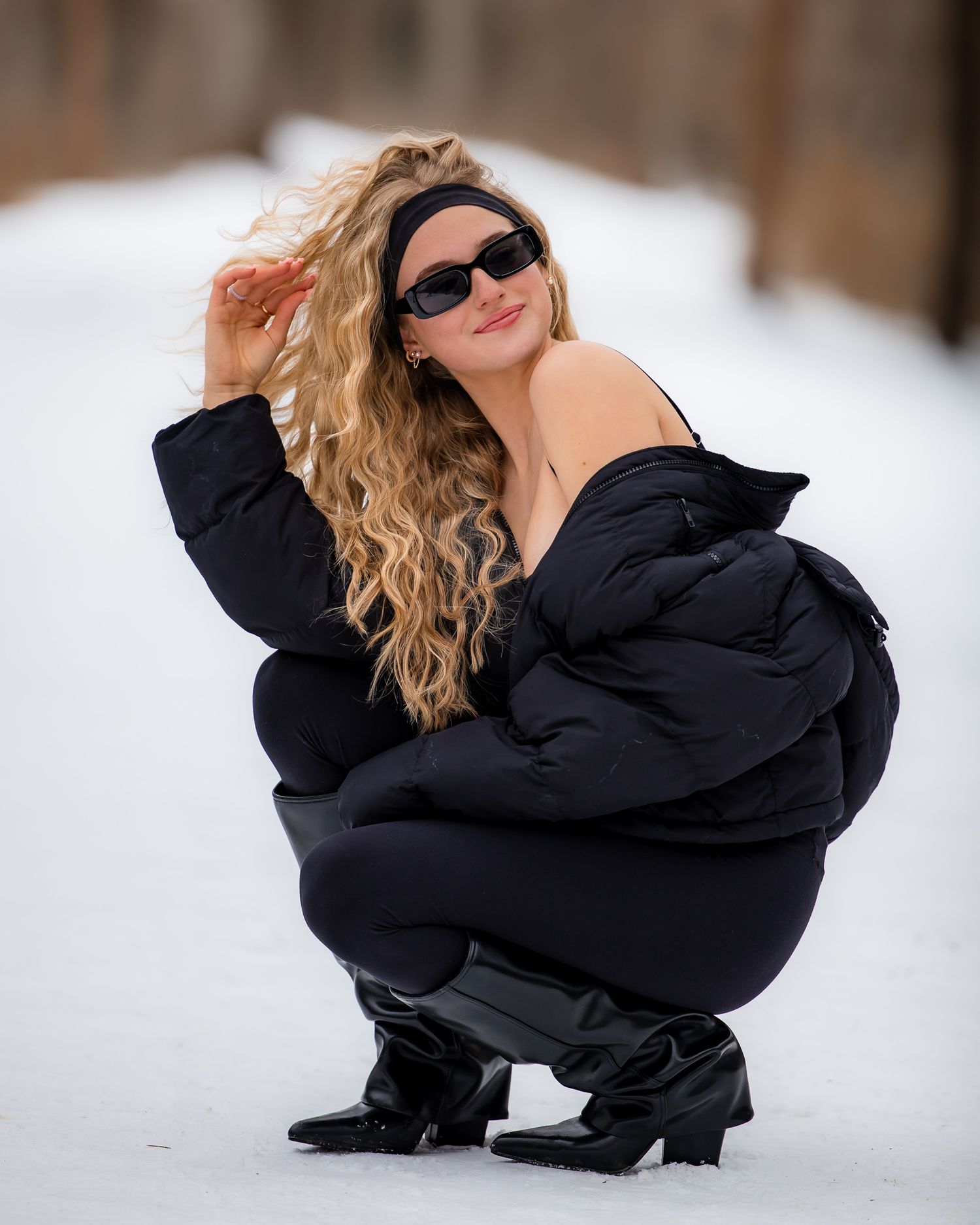 A person in a black winter coat crouches in the snow wearing sunglasses and dark clothing.