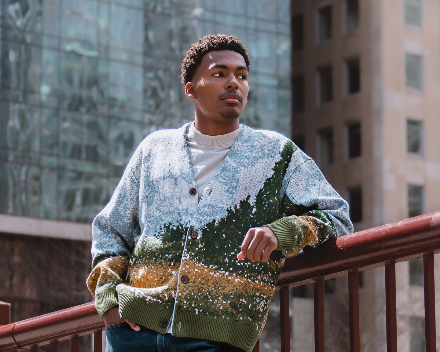 Young man in scenic landscape cardigan sweater leaning on red railing with urban buildings in background.