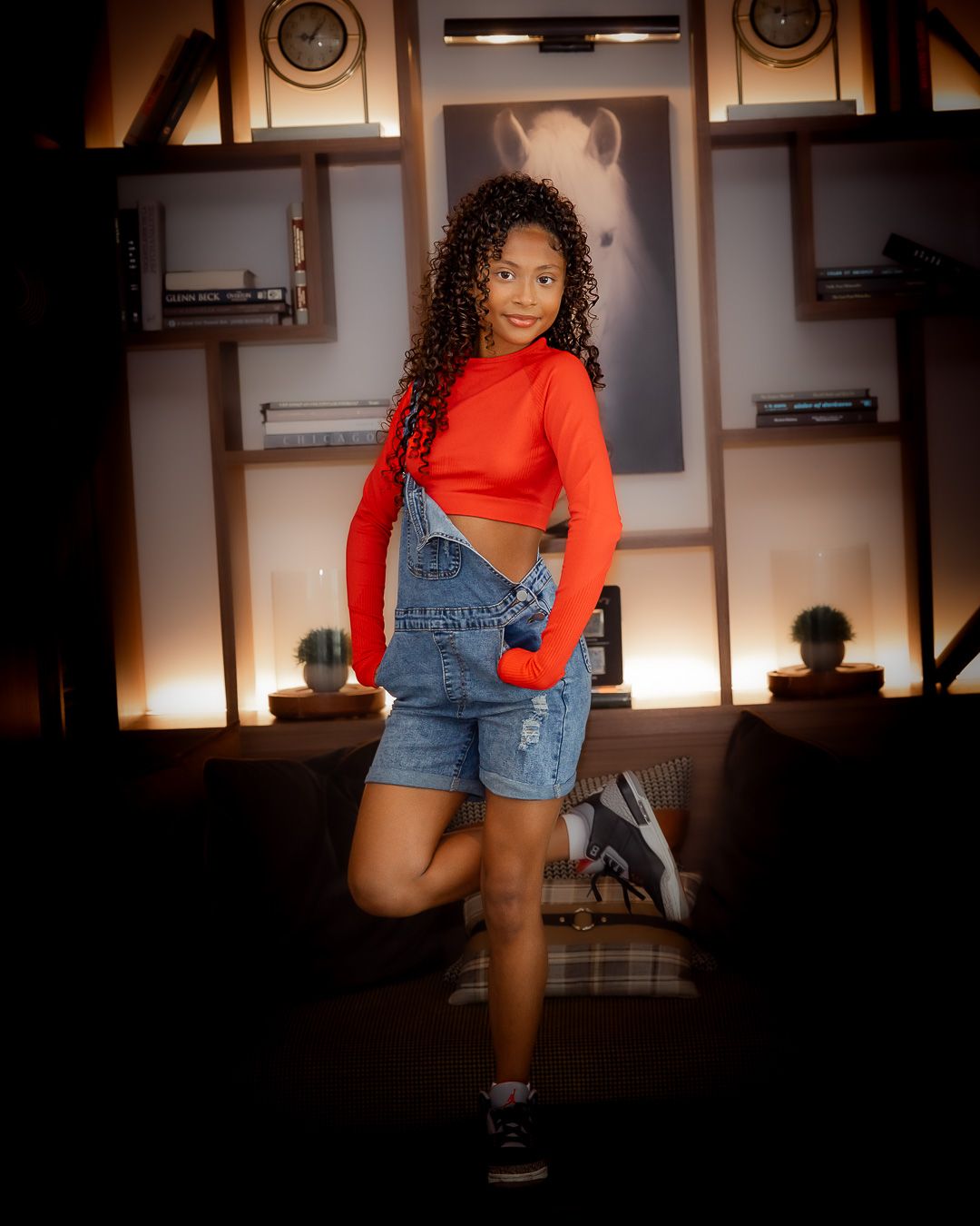 Person in red crop top and denim overalls shorts poses in front of modern shelving display with warm lighting.