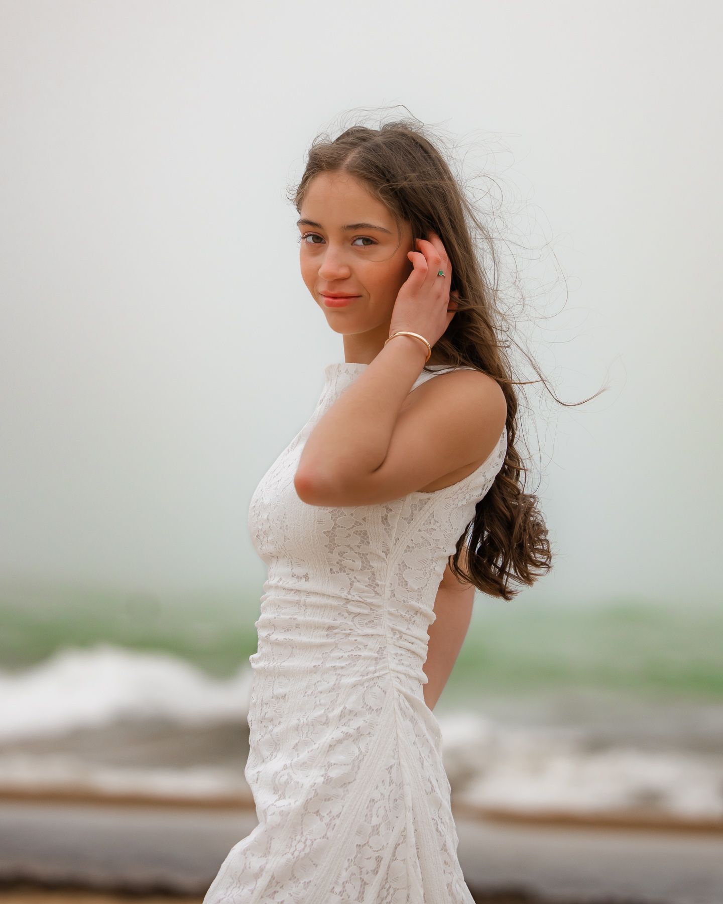 Young woman in white lace dress poses gracefully on a misty beach with wind-blown hair.