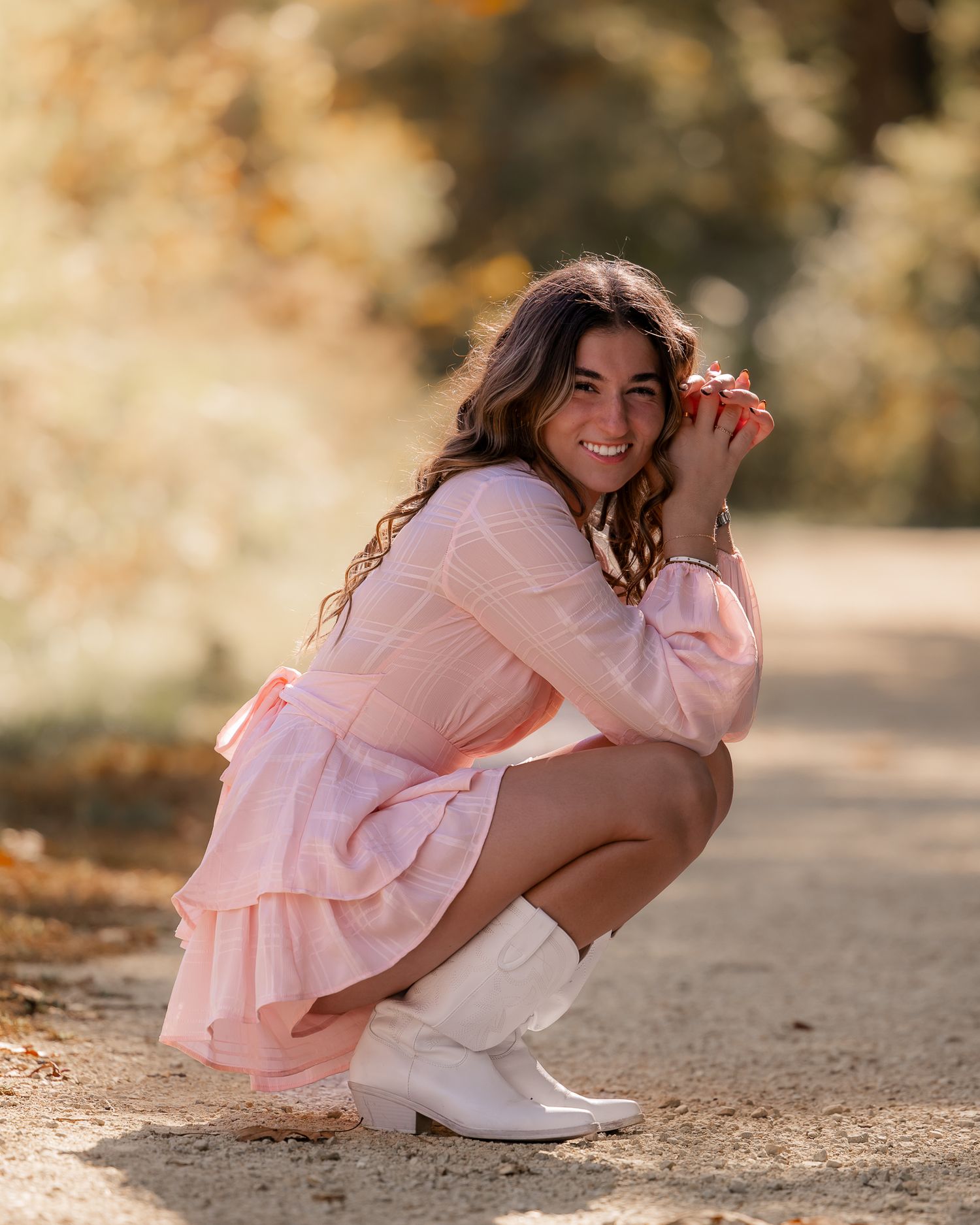 A person in a pink dress and white boots crouches down on a dirt path during a sunny autumn day.