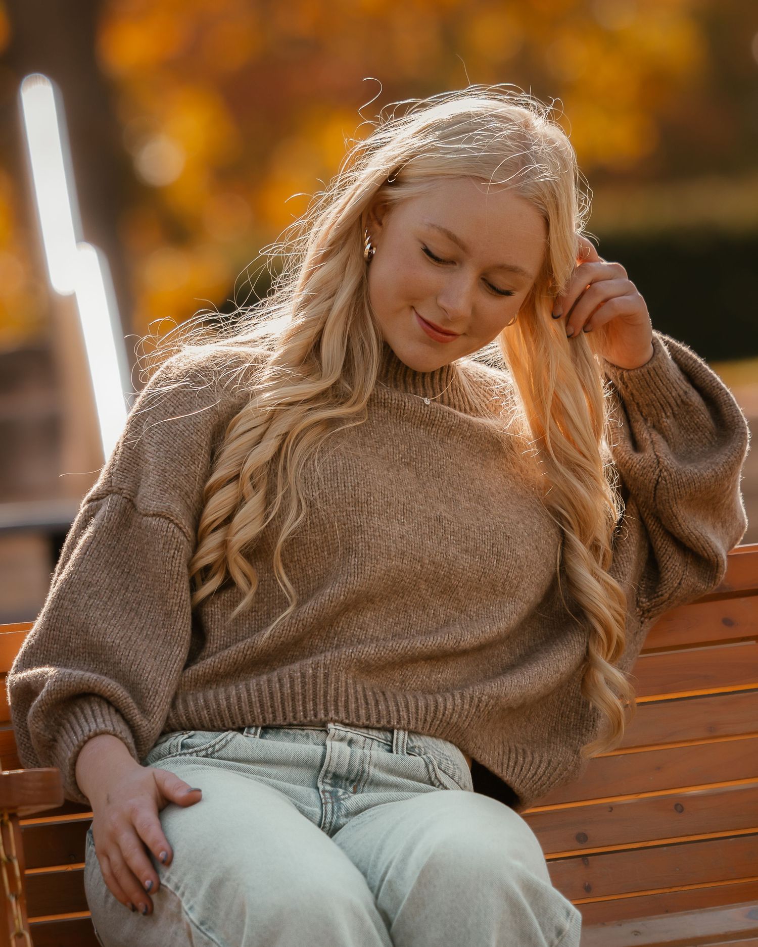 Person in beige sweater sits on wooden bench during autumn with warm golden lighting in background.
