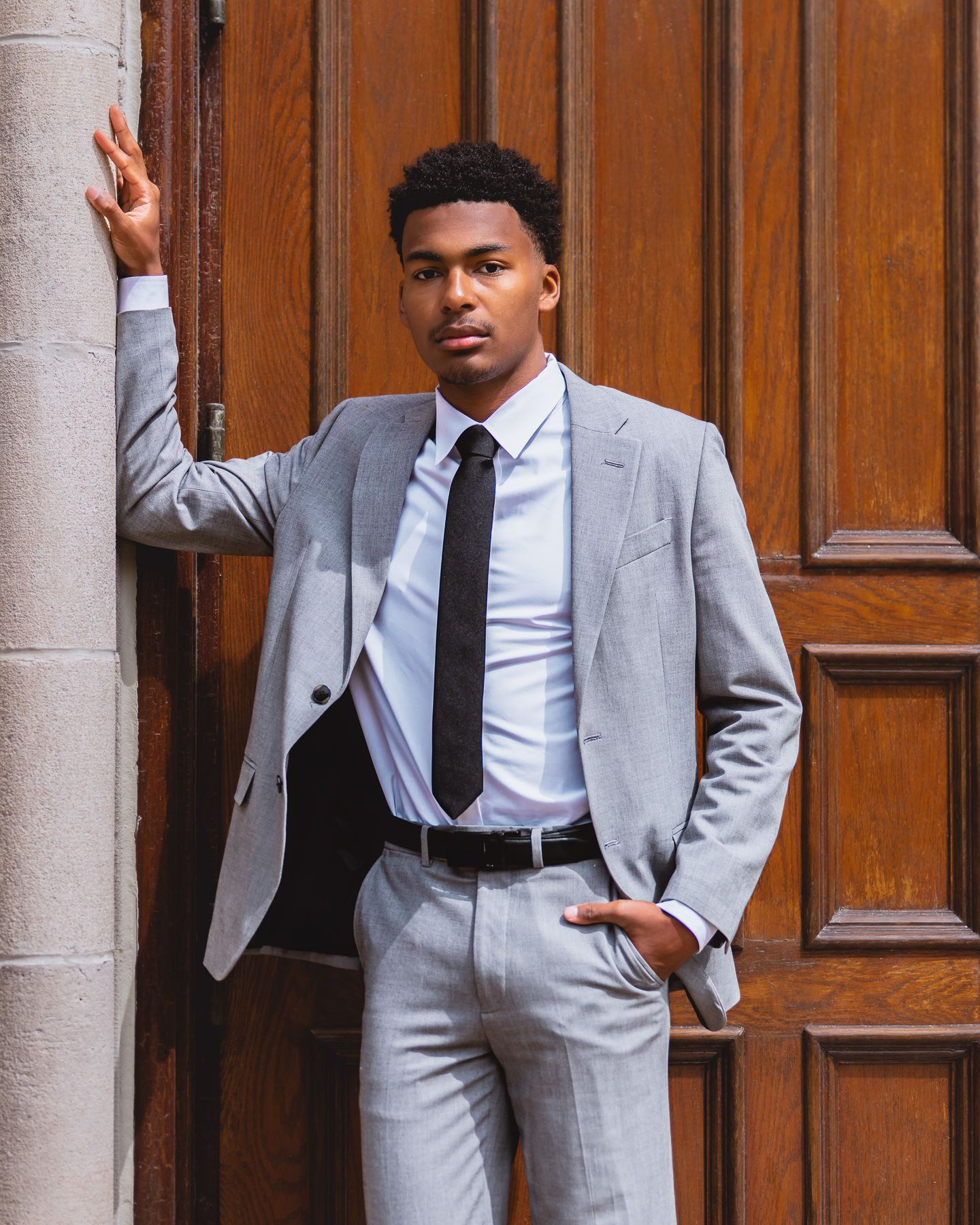 Young man in gray suit and black tie leaning against wooden door in professional portrait photo.