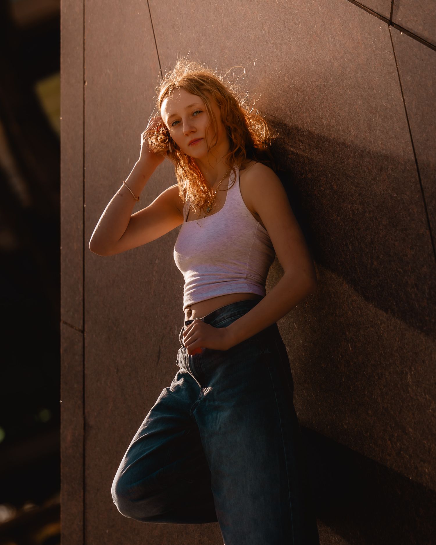 Young woman with red hair in white tank top and jeans leaning against a stone wall in golden hour light.
