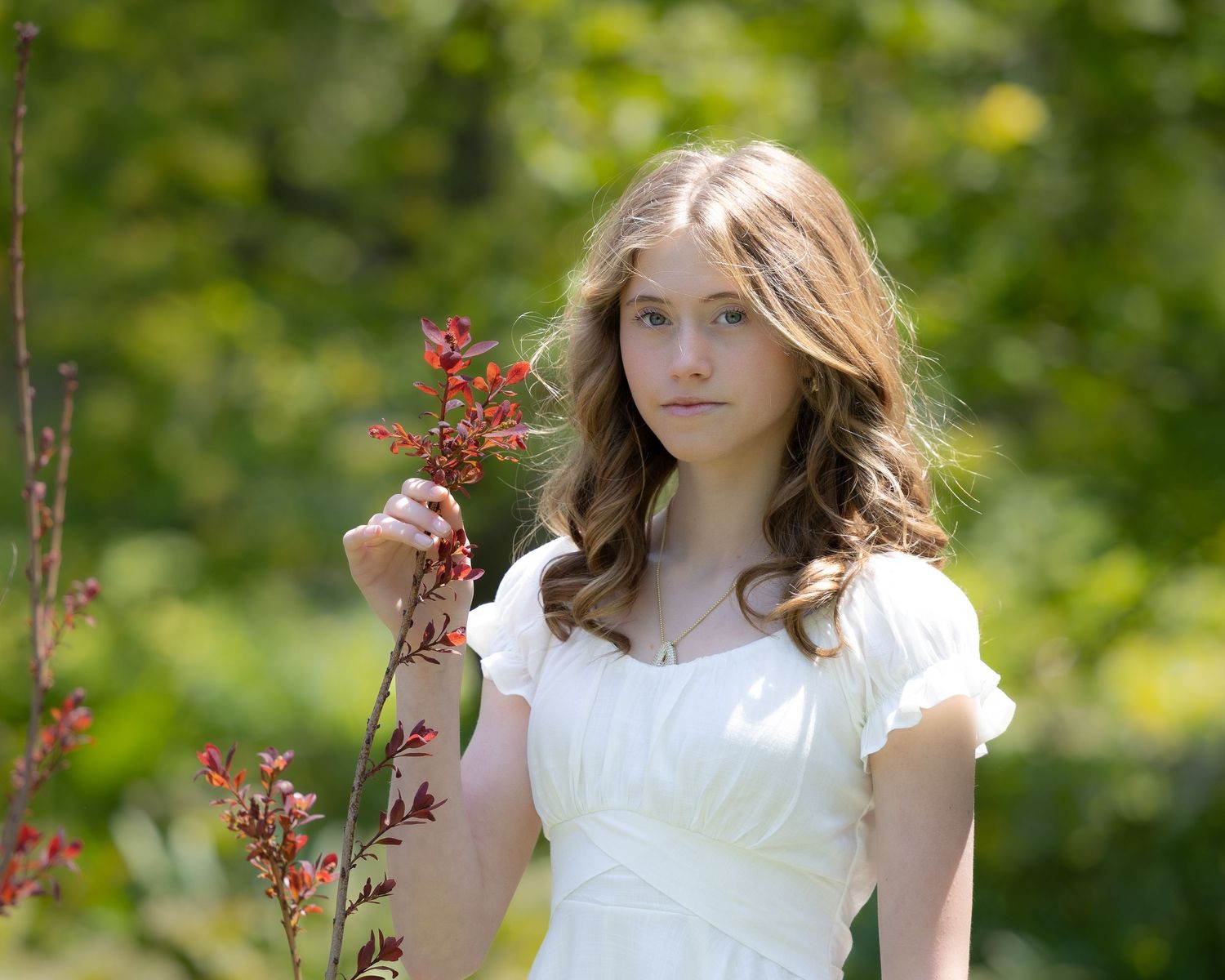 Woman in white dress stands in vibrant green garden holding red flowering branch on sunny summer day.