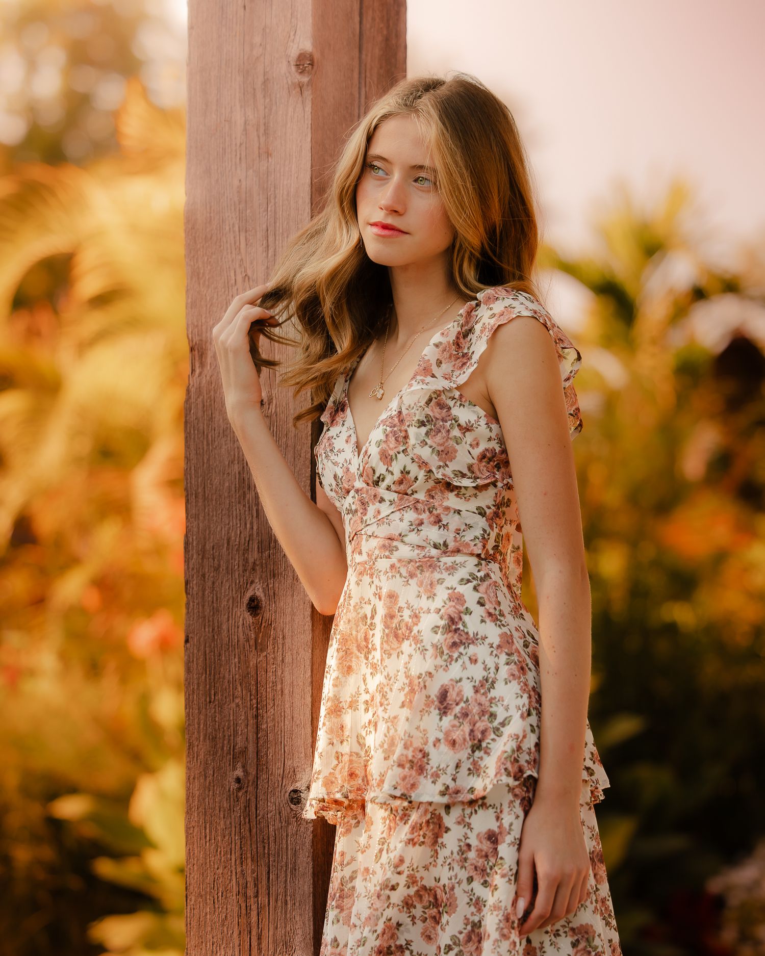 Woman in floral dress poses against wooden post during golden hour with autumn foliage.
