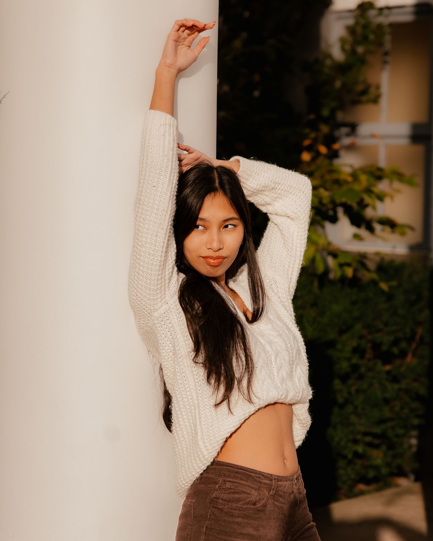 Woman in white knit sweater and brown pants poses against white wall with greenery in background.