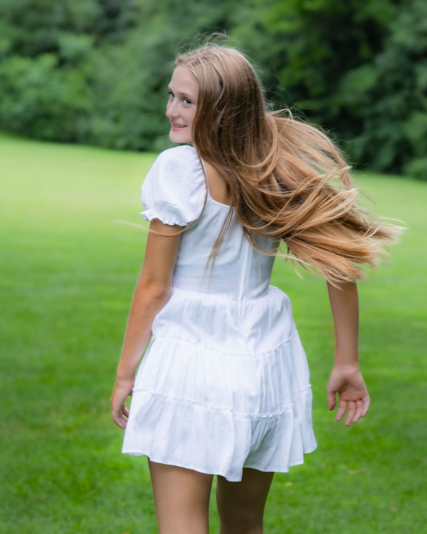 Young girl in white dress twirling outdoors on green lawn, hair flowing, smiling over shoulder.