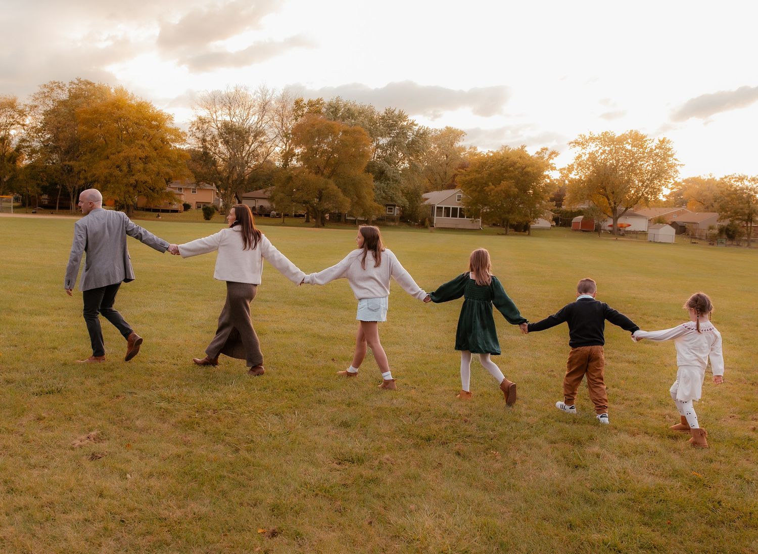 Family of six holding hands walking across a green field at sunset with autumn trees in the background.