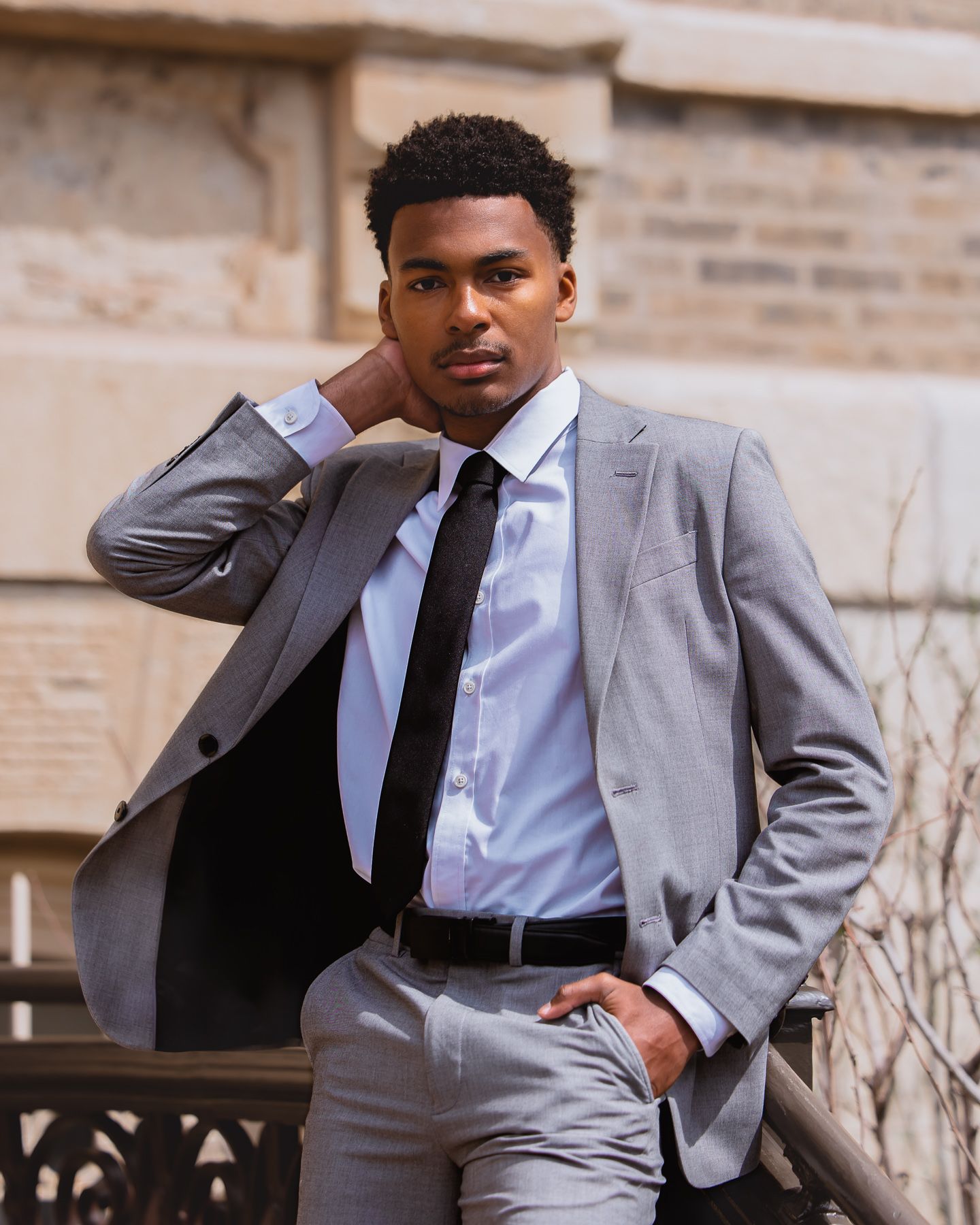 Young man in gray suit and black tie posing confidently outdoors near stone architecture.