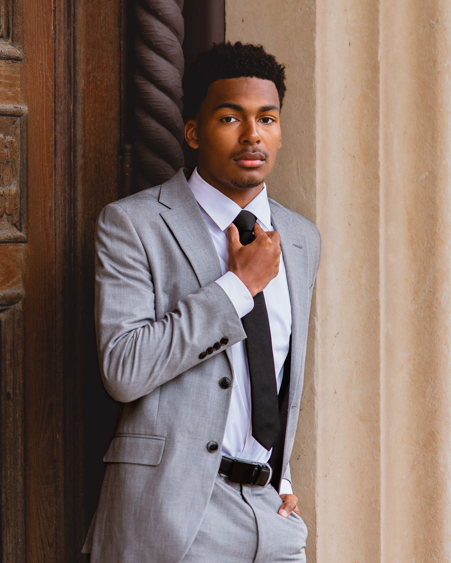 Young man in gray suit adjusting black tie, leaning against stone column outdoors.