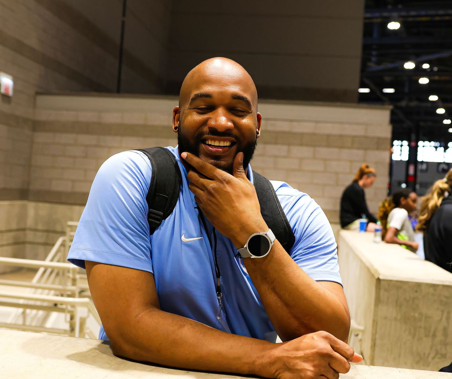 Person in light blue t-shirt smiles while sitting at table in convention center setting.