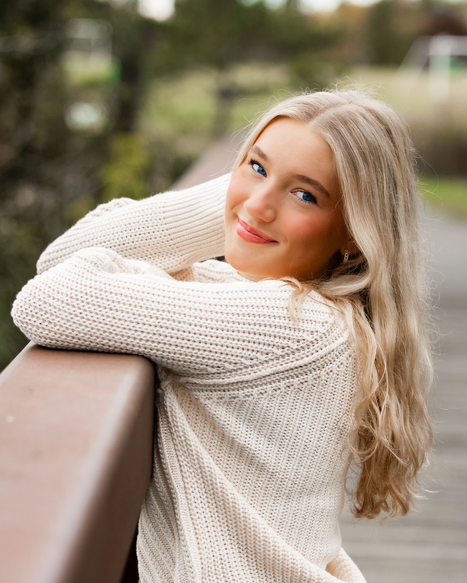 A person in a cream knit sweater poses against wooden railings.