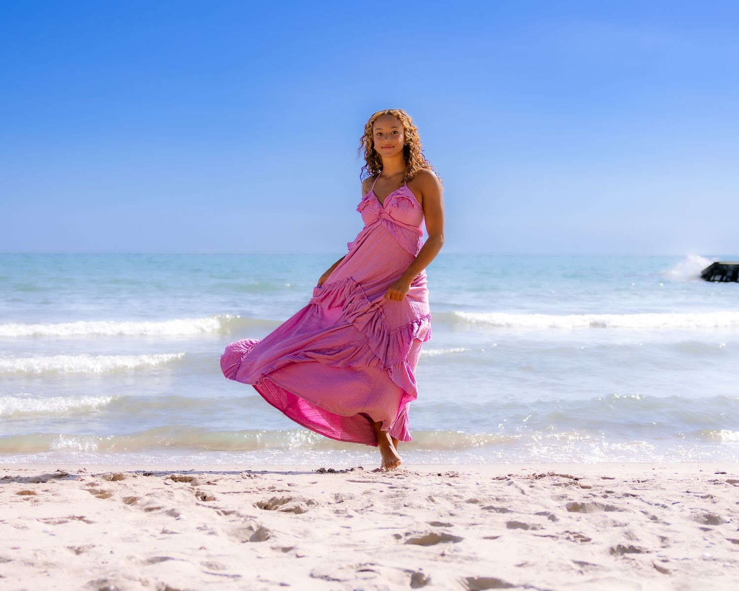 Woman in flowing pink dress walking on a sunny beach with blue ocean waves in the background.