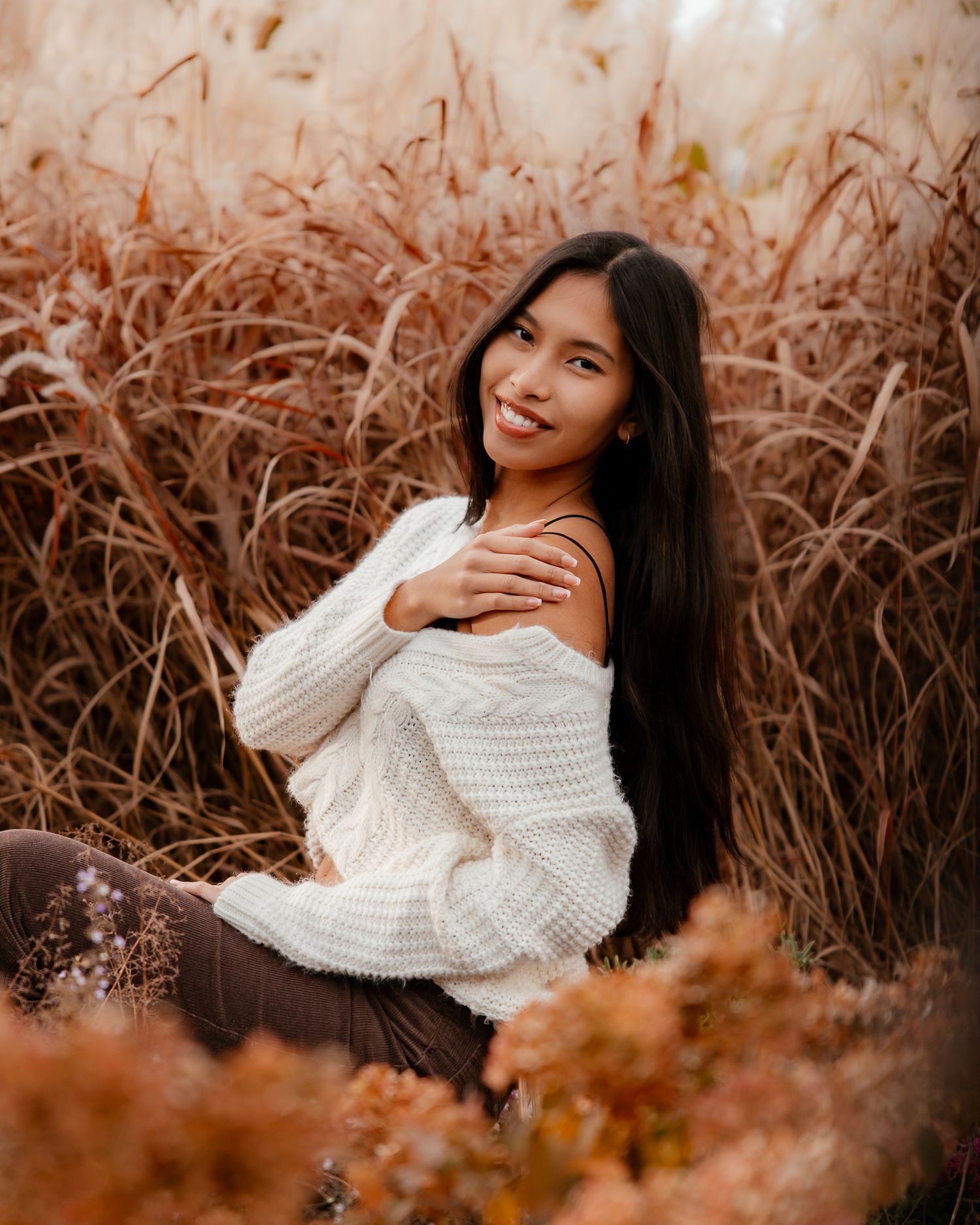A person in a white sweater poses in a field of tall brown grass during autumn at sunset.