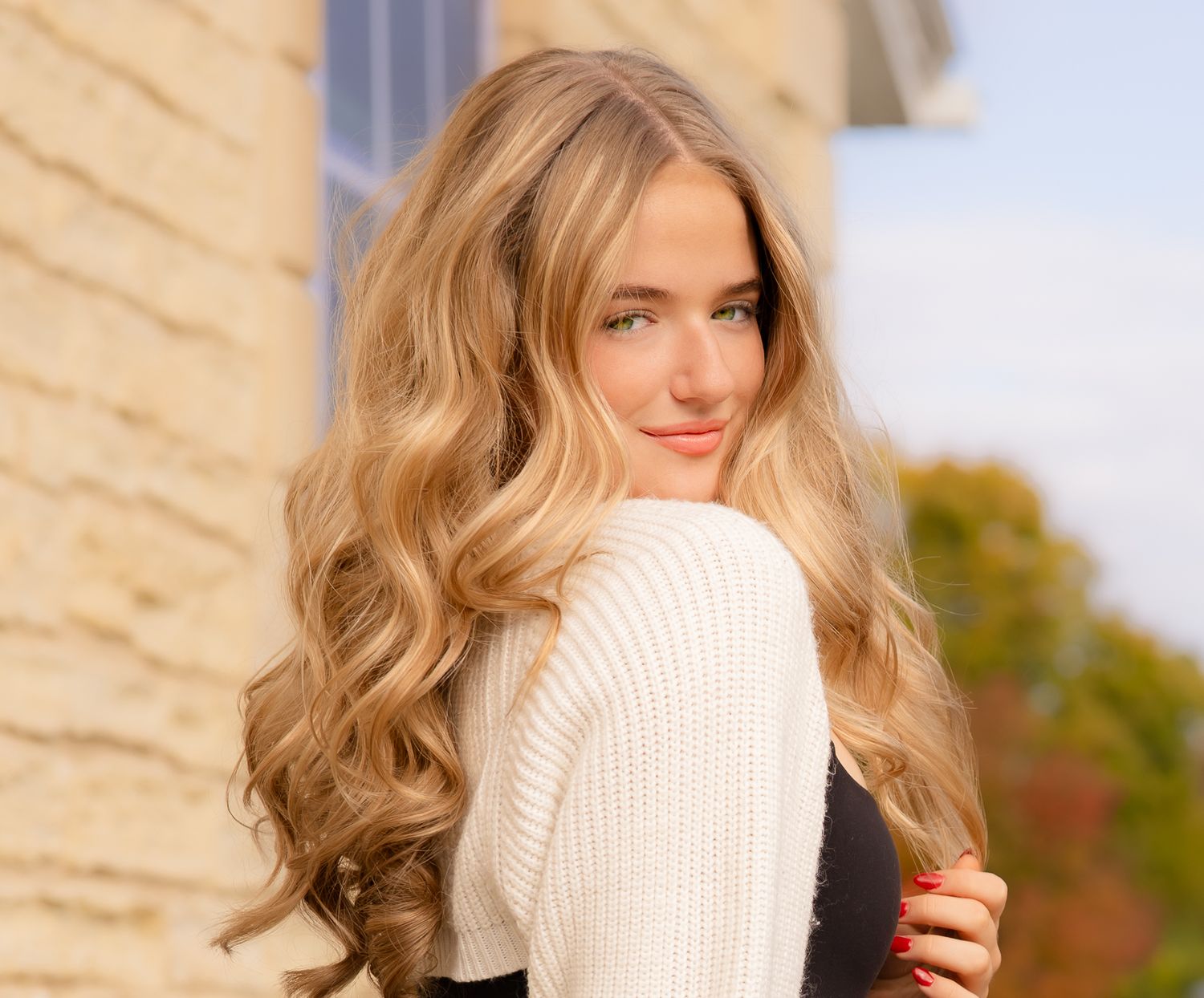 Portrait of a young woman in a cream sweater with long wavy blonde hair against a light background.