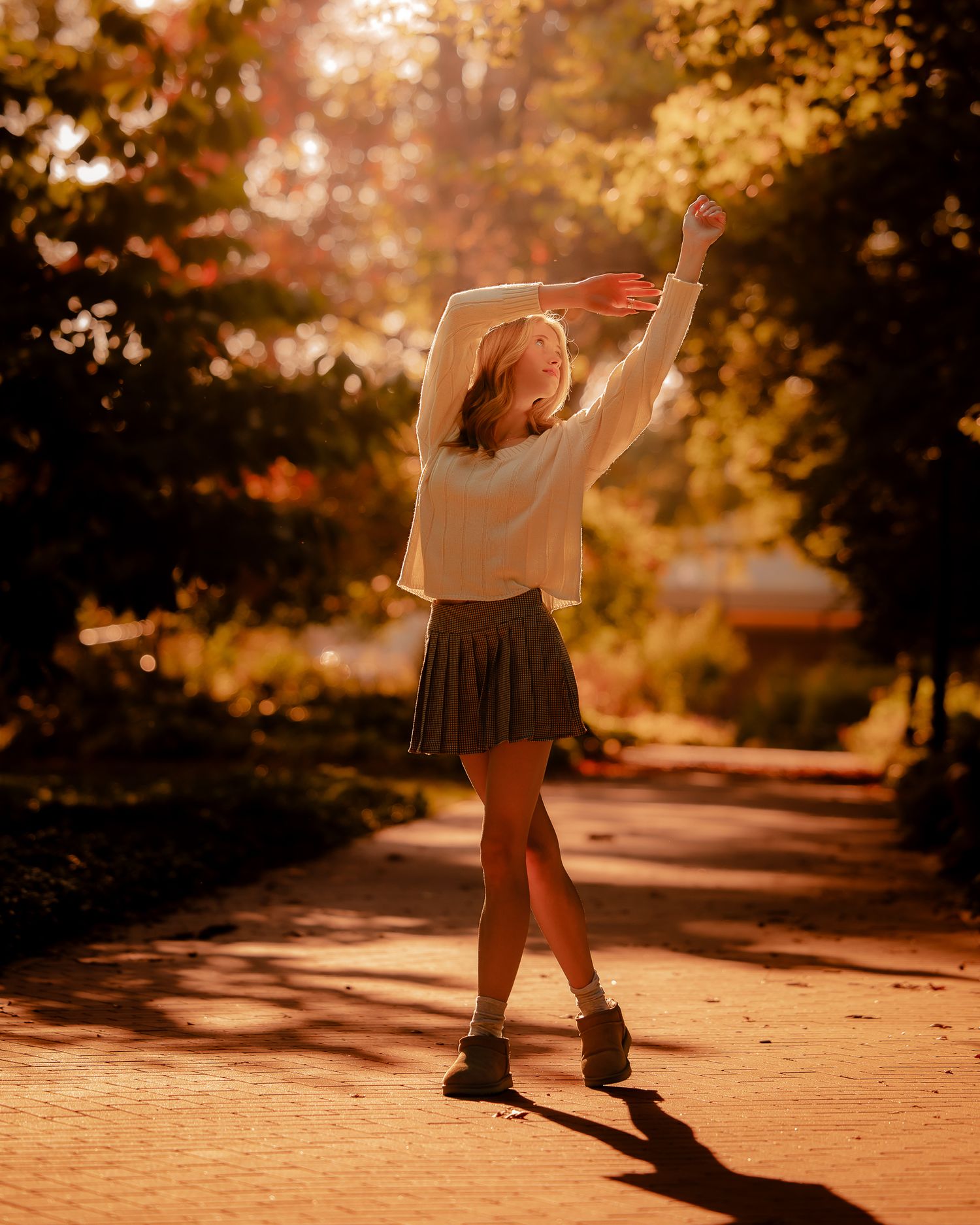 A young woman in a white top and dark pleated skirt dances joyfully on a sunlit path surrounded by autumn trees.