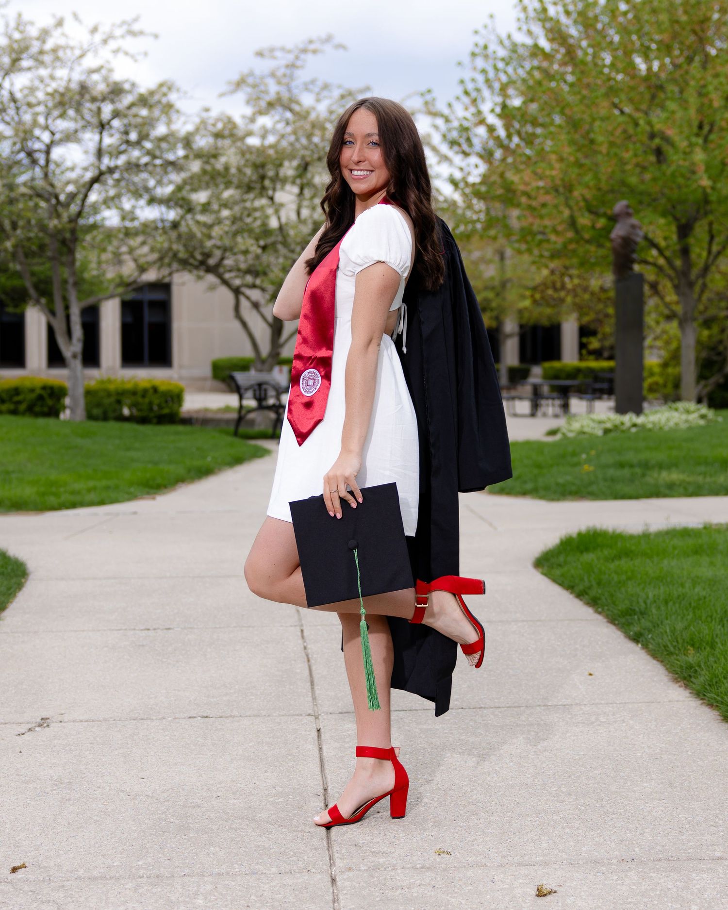 Graduate in red heels and academic gown poses on campus sidewalk.