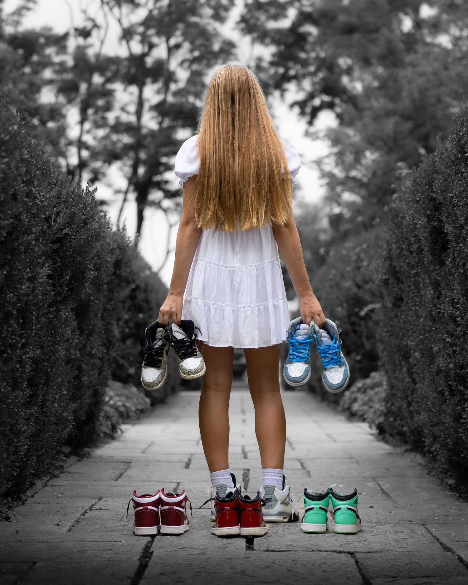 A person in a white dress holding pairs of colorful sneakers while standing on a path lined with green hedges.