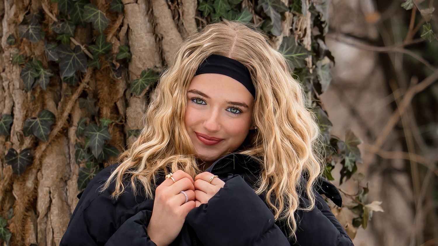 Blonde woman in black winter coat smiles while leaning against tree trunk with ivy in outdoor setting.