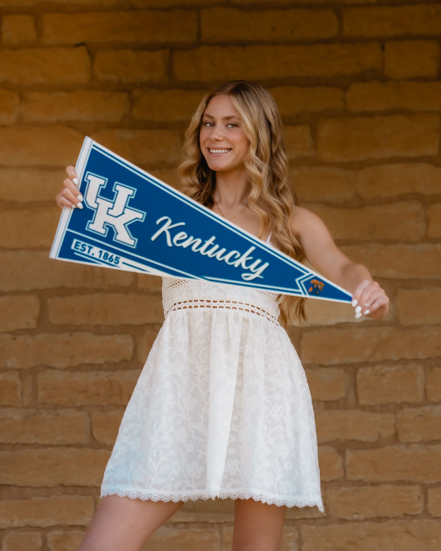 Smiling blonde woman in white lace dress holding a University of Kentucky blue pennant flag outdoors.