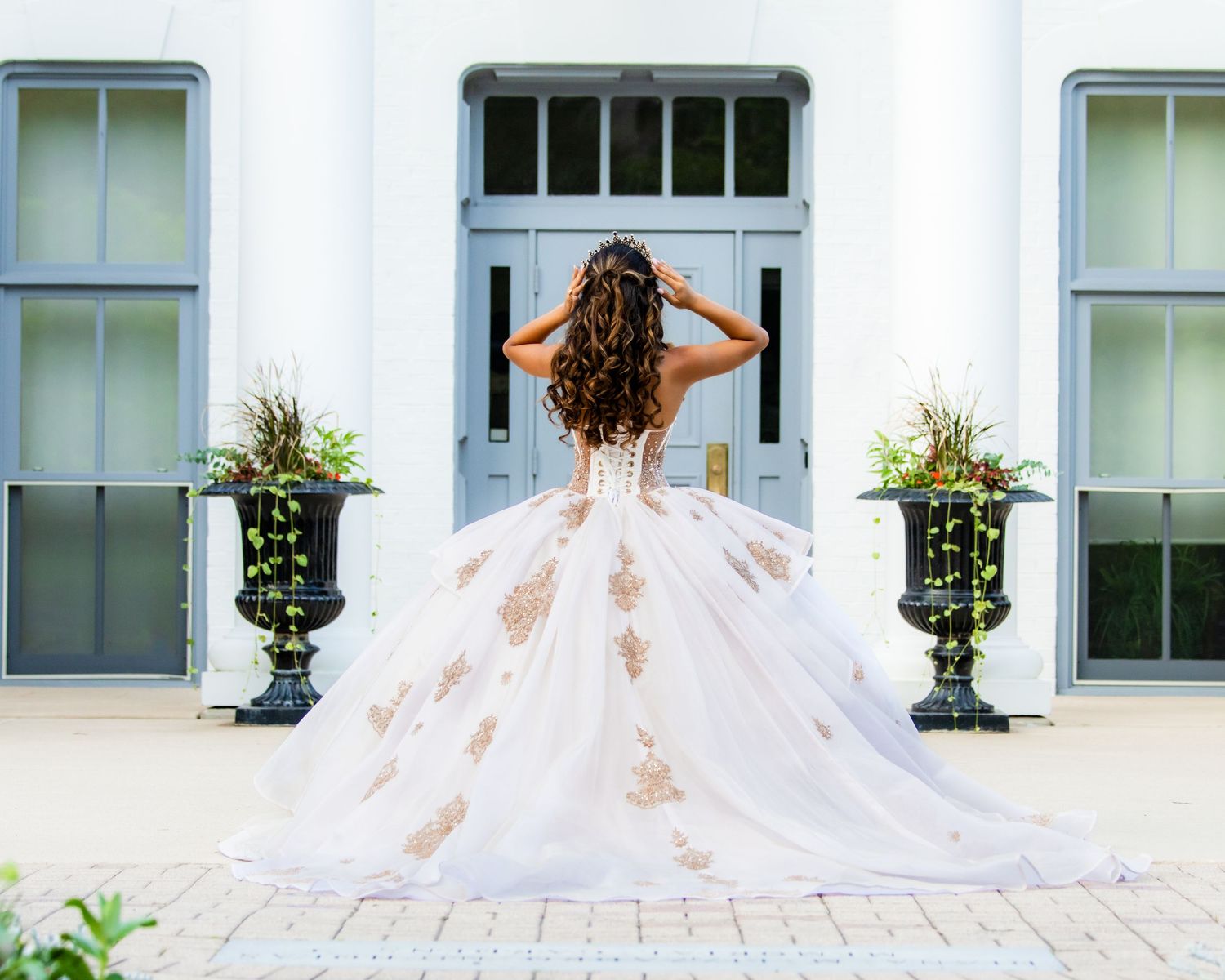 A bride in an ornate white flowing ballgown stands before a light blue door with potted plants on either side.