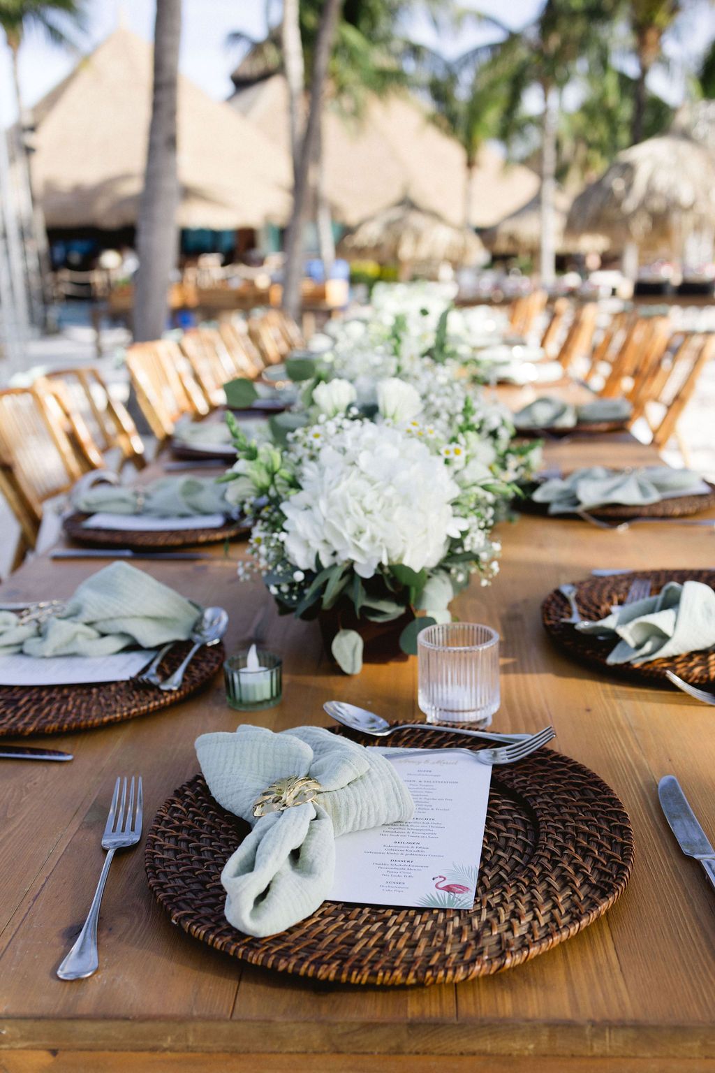 Elegant beach wedding reception table set with wicker chargers, white florals, and sage napkins.